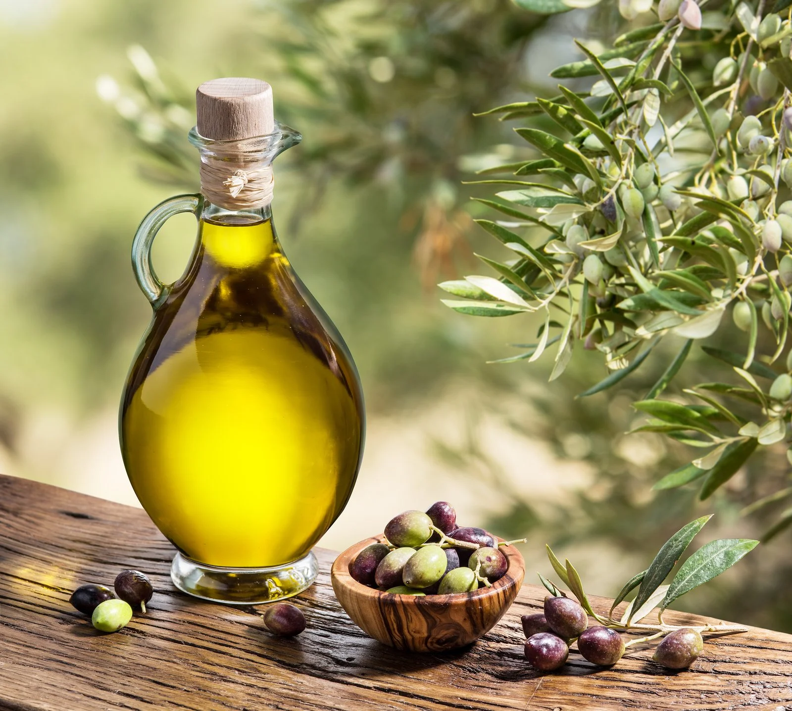 A glass bottle of olive oil with a wooden cap, a small bowl of green and purple olives, and olive branches with leaves and unripe olives on a wooden surface outdoors.