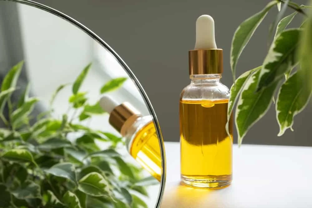 A glass dropper bottle filled with vitamin E oil, placed on a white surface near a mirror and green plant leaves.