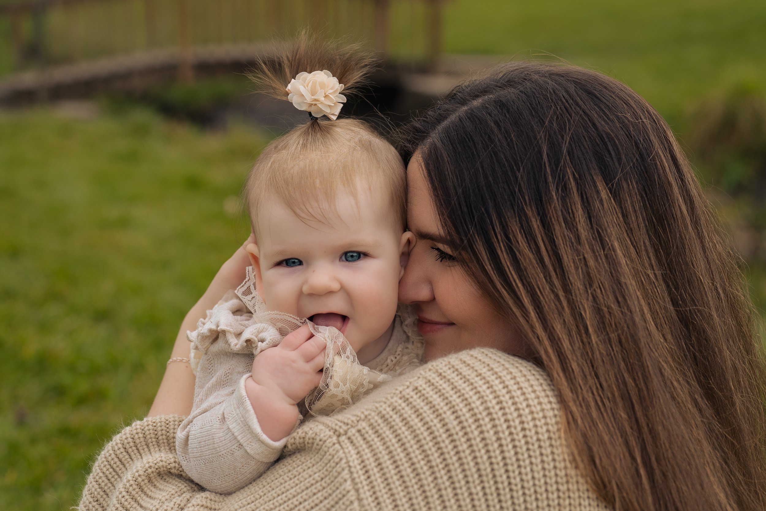A woman holding a young girl outdoors in a grassy area, with a fence and trees in the background. The girl has blonde hair with a flower accessory and is licking her fingers, while the woman is smiling and resting her face against her head.
