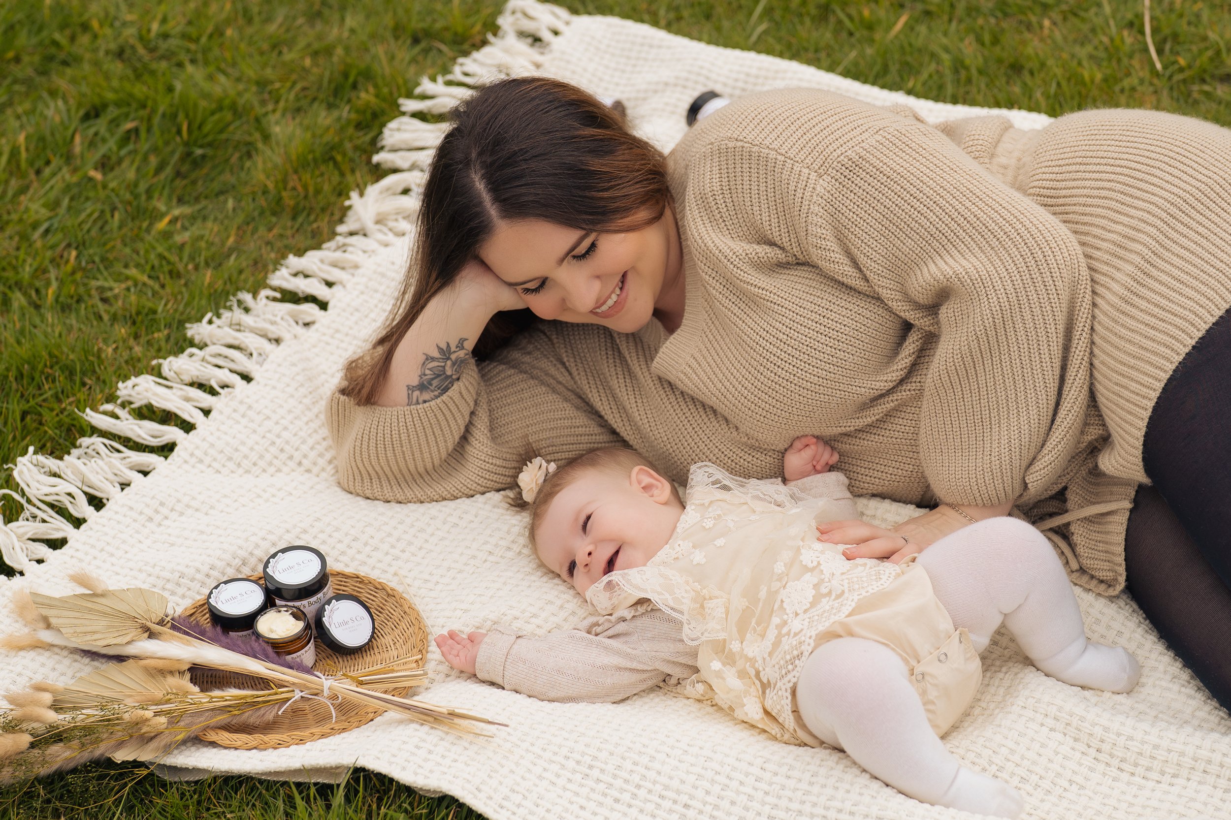 A woman and a baby girl lying on a blanket on the grass, smiling and looking at each other. The woman is resting her head on her hand, wearing a beige sweater, and has a tattoo on her arm. The baby is dressed in a cream-colored dress with lace details and white tights. There are skincare products and dried flowers on a woven tray beside them.