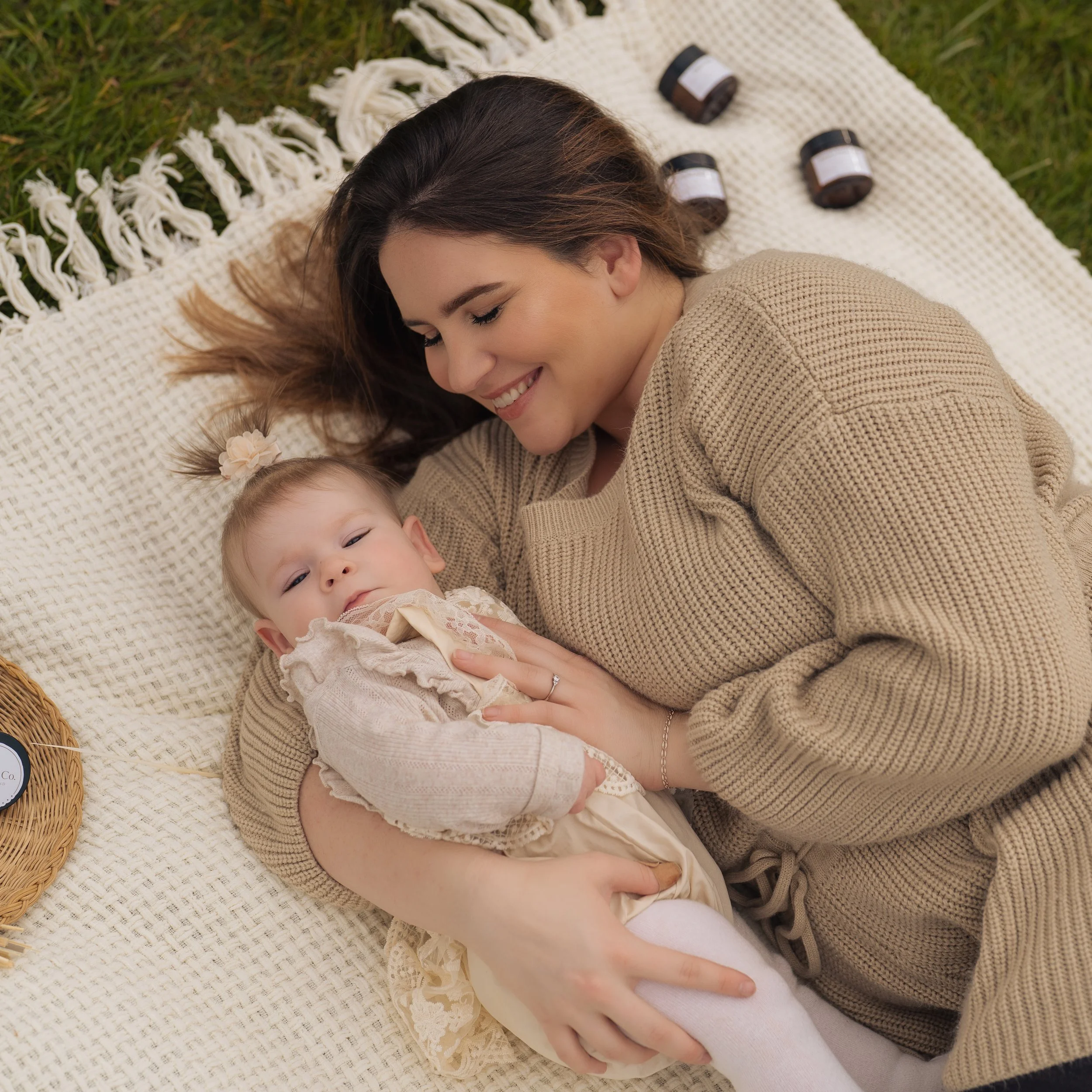 A woman and a young girl lying on a blanket outdoors, smiling and enjoying each other's company, with jars of natural, tallow-based skincare by Little S Co. and a basket nearby.
