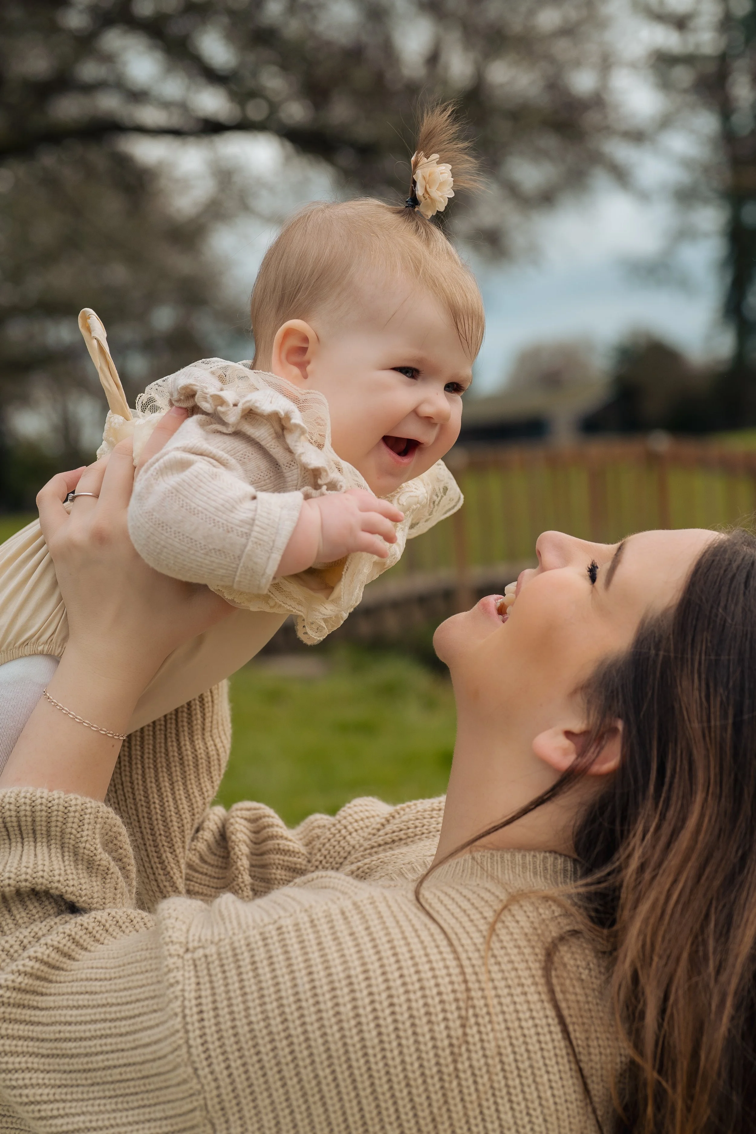 A woman lifting a smiling baby girl with short light hair styled in a top knot with a flower accessory outdoors. The woman has long brown hair and is wearing a beige sweater. They are enjoying a joyful moment in a grassy area with trees and a wooden fence in the background.