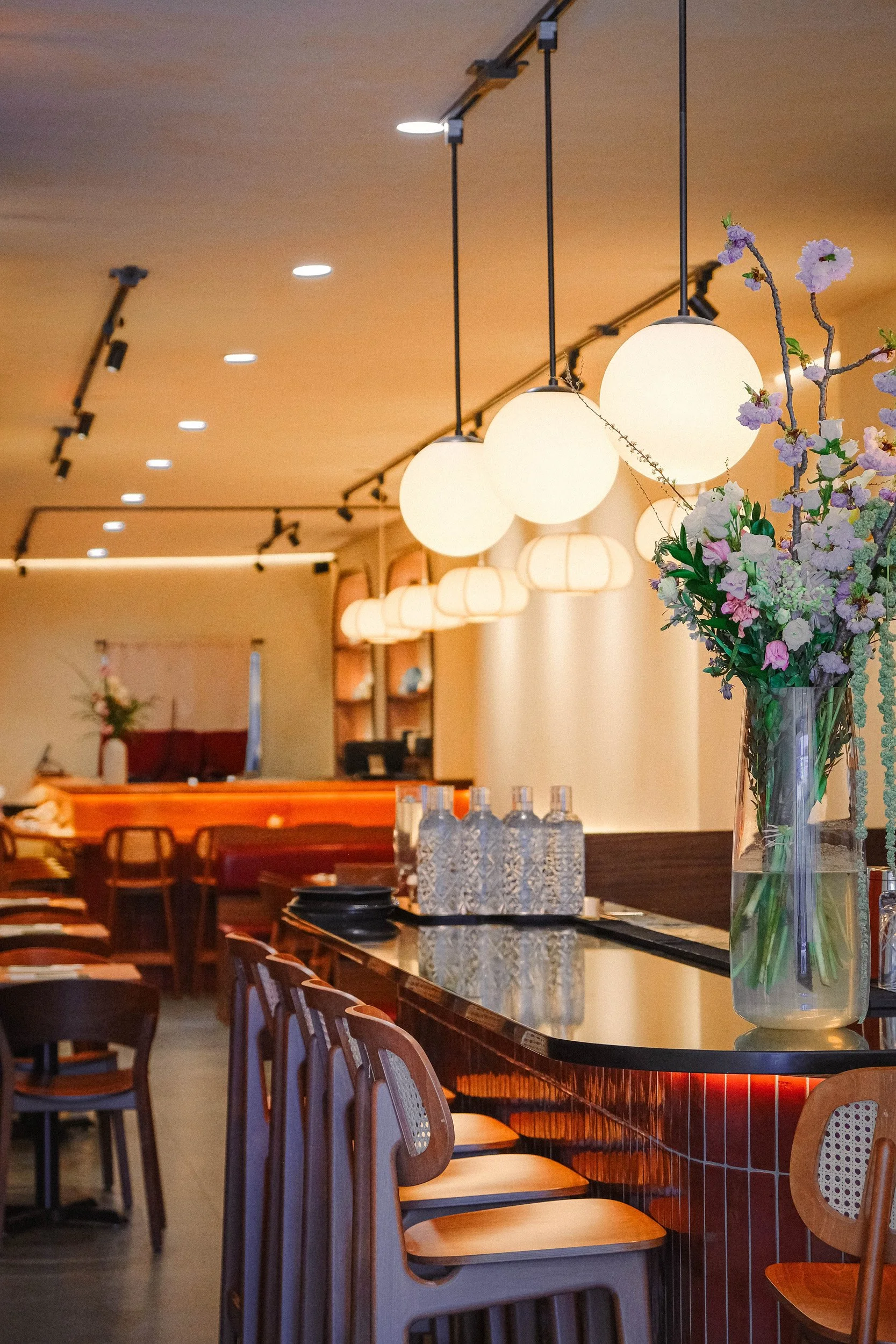 Interior of a restaurant with a bar counter, wooden chairs, and hanging globe pendant lights. A large vase of pink and purple flowers is on the counter. The background shows a small seating area with cushions and a partition.