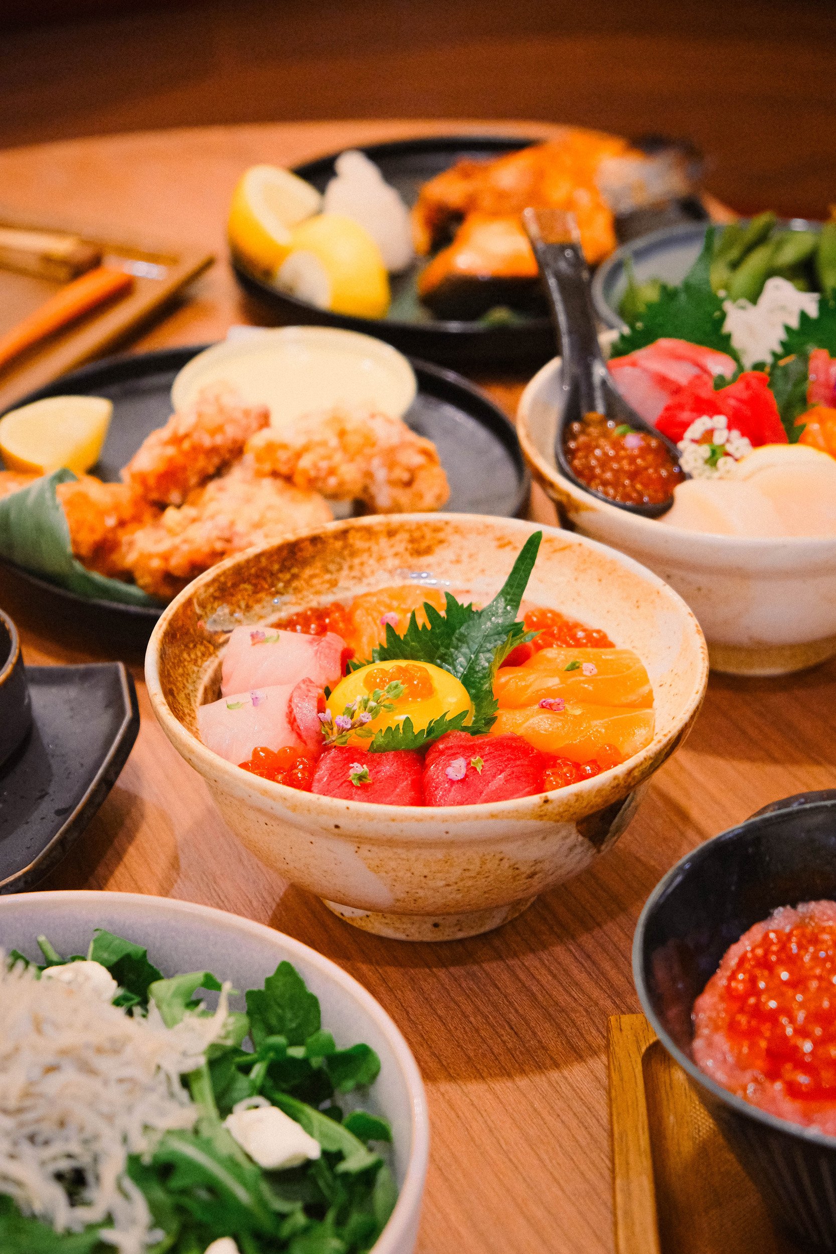 Assorted Japanese dishes including rice bowls, sashimi, salads, and seafood with lemon wedges and condiments on a wooden table.
