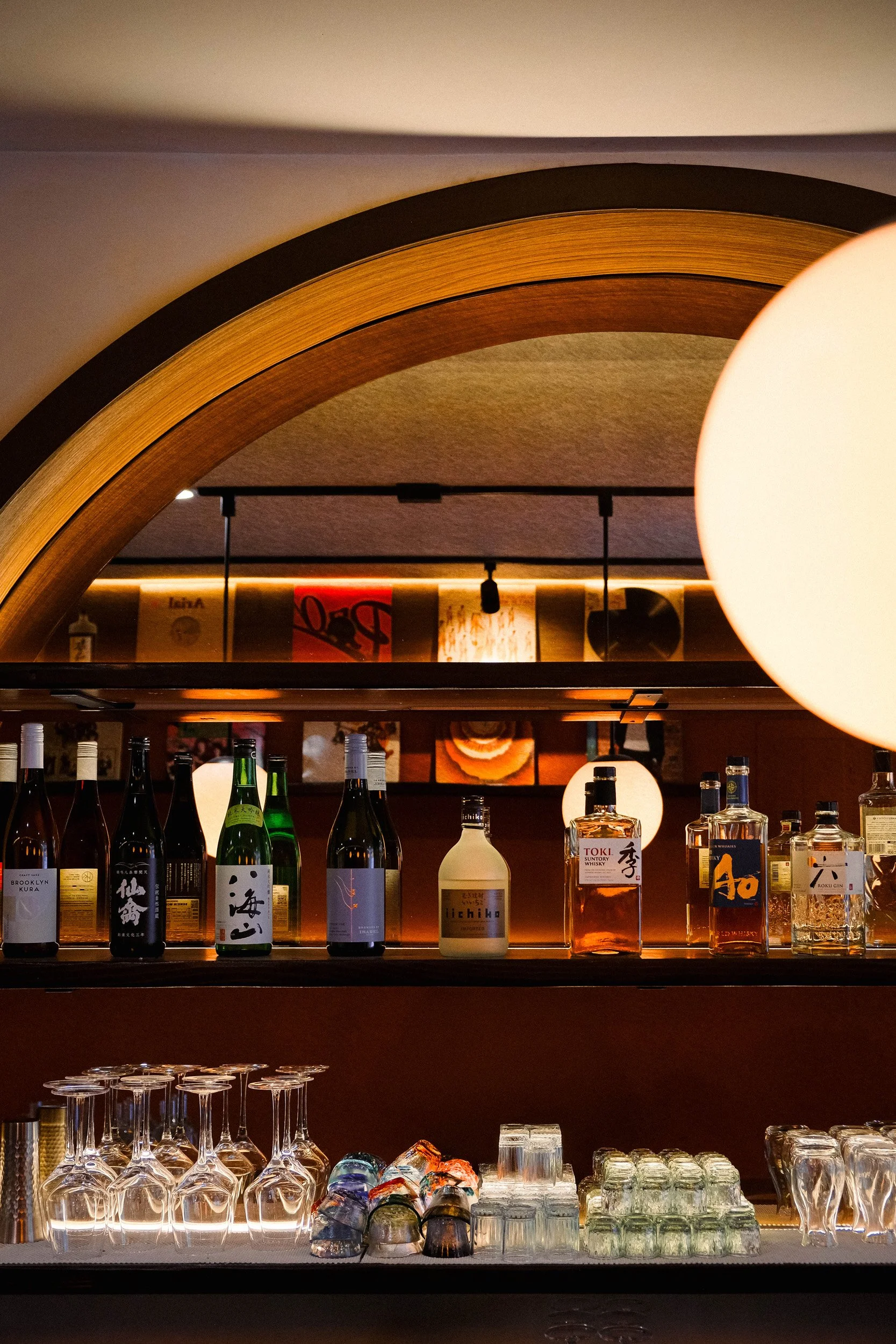 Bar shelf with various liquor bottles, upside-down wine glasses, shot glasses, and ice cubes, with warm lighting and a decorative arch.