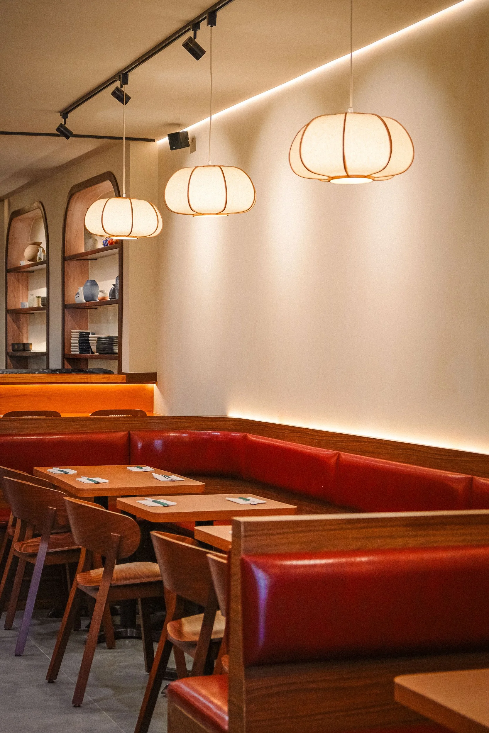 Interior view of a restaurant dining area with wooden tables, chairs, a curved red bench, hanging lantern-style lights, and shelves with decorative items on a wall.