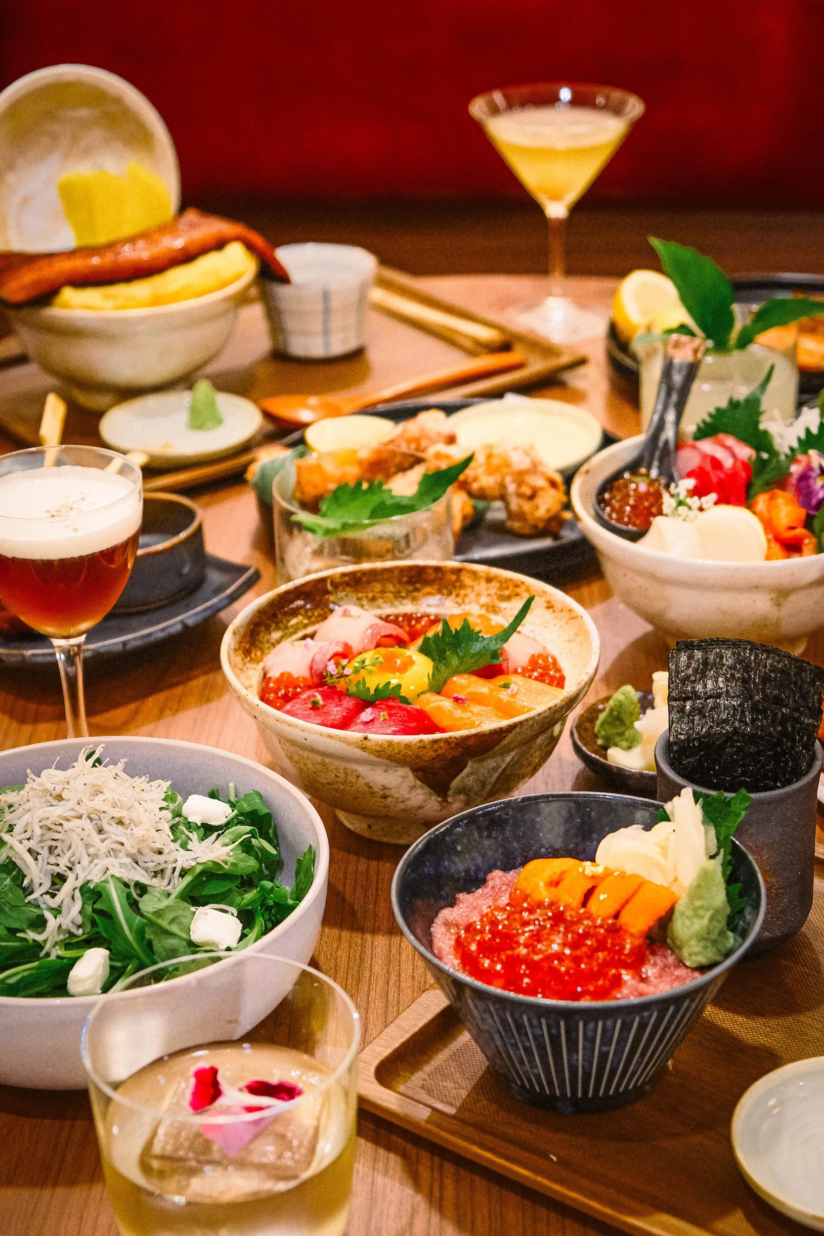 A table filled with various dishes including rice bowls, sashimi, salads, small plates with sauces, and drinks, indicating a Japanese or Asian cuisine meal.