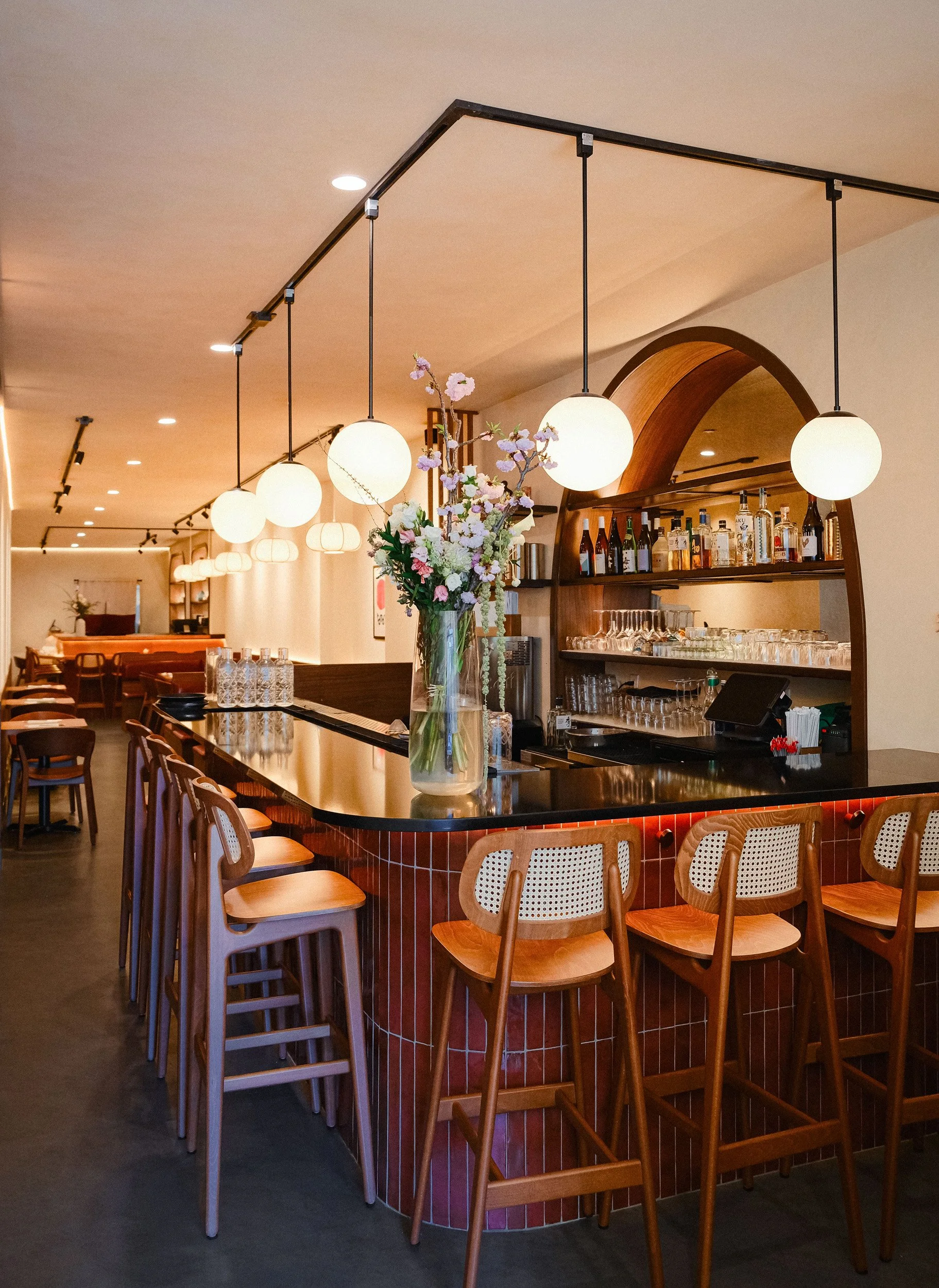 An interior of a modern bar with a curved black bar counter, wooden chairs, and hanging round white pendant lights. A floral arrangement with purple and white flowers is on the bar counter. Shelves behind the bar display bottles of alcohol and glassw