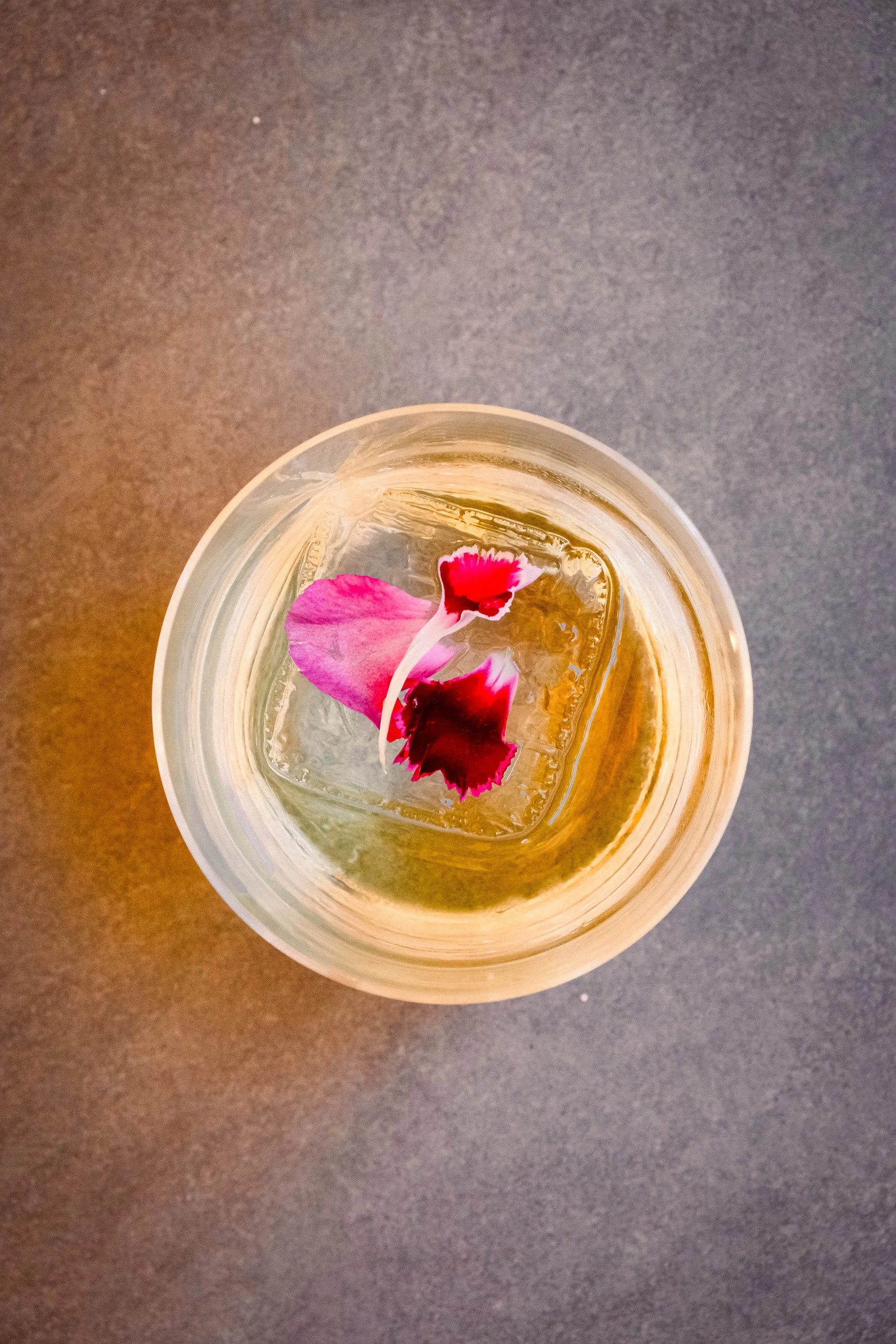 A clear glass of water with a pink and red flower petal floating on top, placed on a dark, flat surface.