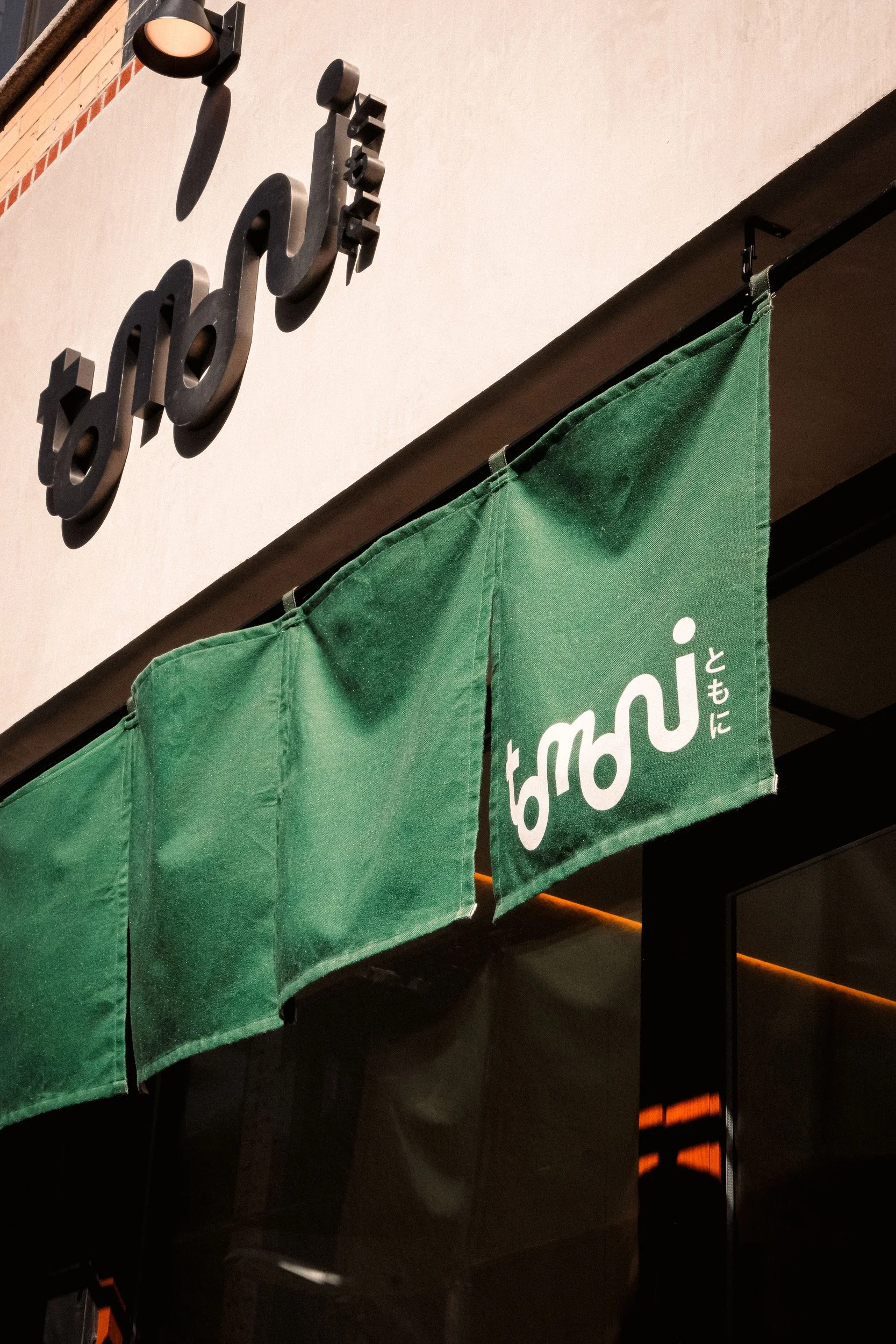 Part of a green fabric curtain or banner with white text, hanging from a black rod, with a dark ceiling and wall behind it. A black logo or sign made of metal with stylized letters or symbols is mounted on the wall above the curtain.