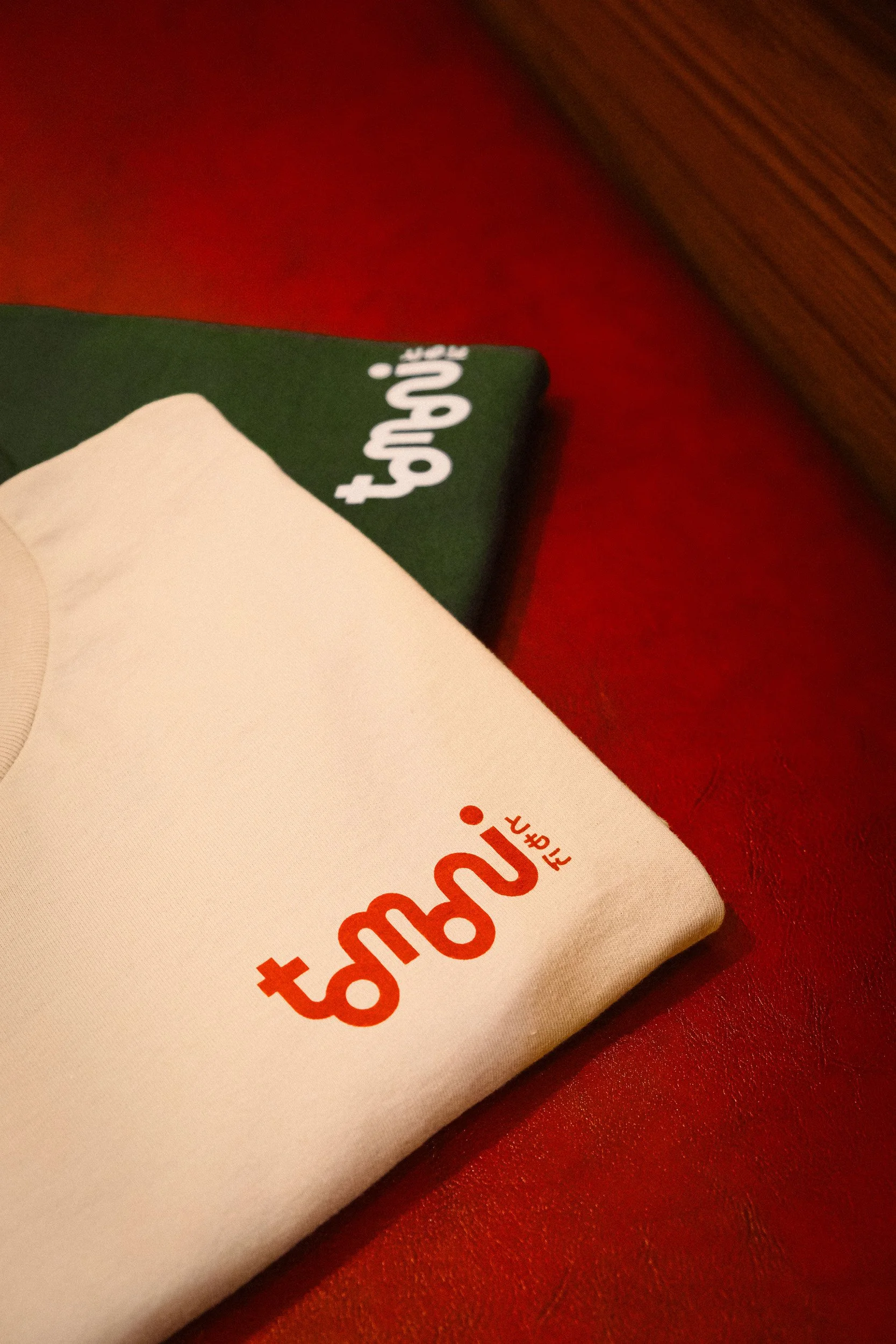 Close-up of a tan t-shirt with red and orange text resting on a red surface, with partially visible green and white t-shirt in the background.