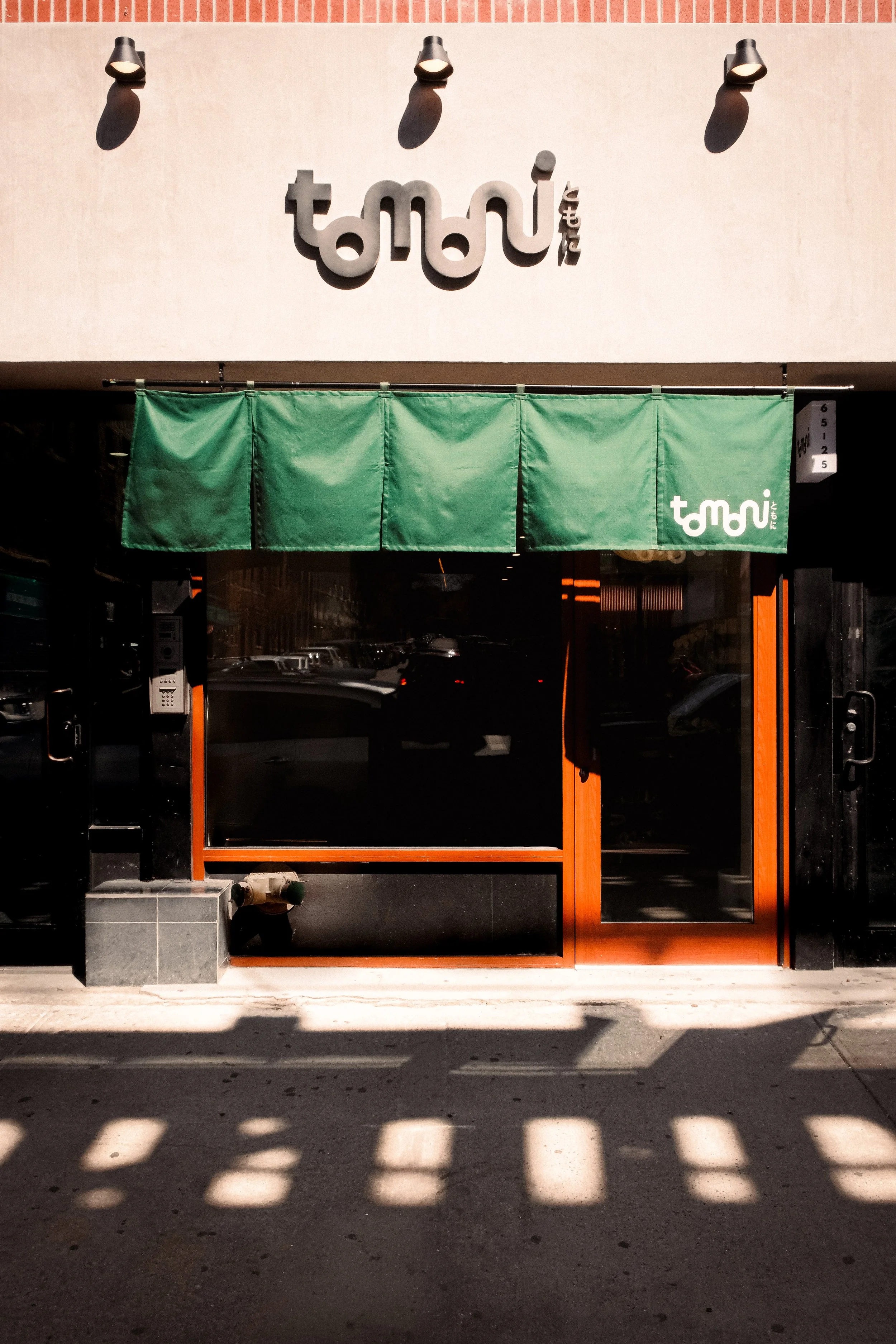 Storefront with Japanese text, green fabric banner, glass door, and orange frame, with reflections and shadows on the sidewalk.