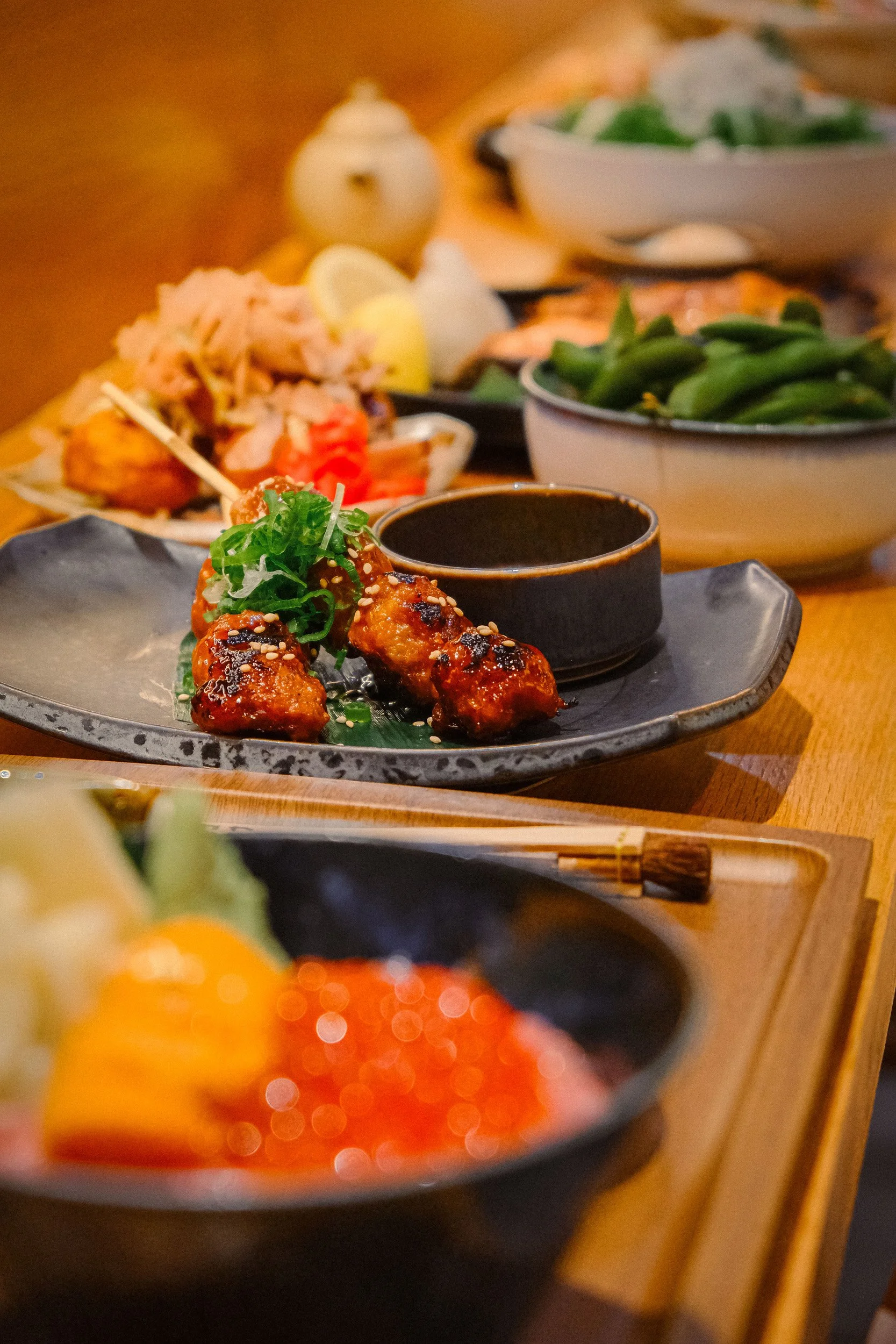 Close-up of a Japanese dish with glazed grilled chicken meatballs garnished with green onions, served with dipping sauce on a black plate, blurred background of other dishes including salad, vegetables, lemon wedge, and chopsticks.