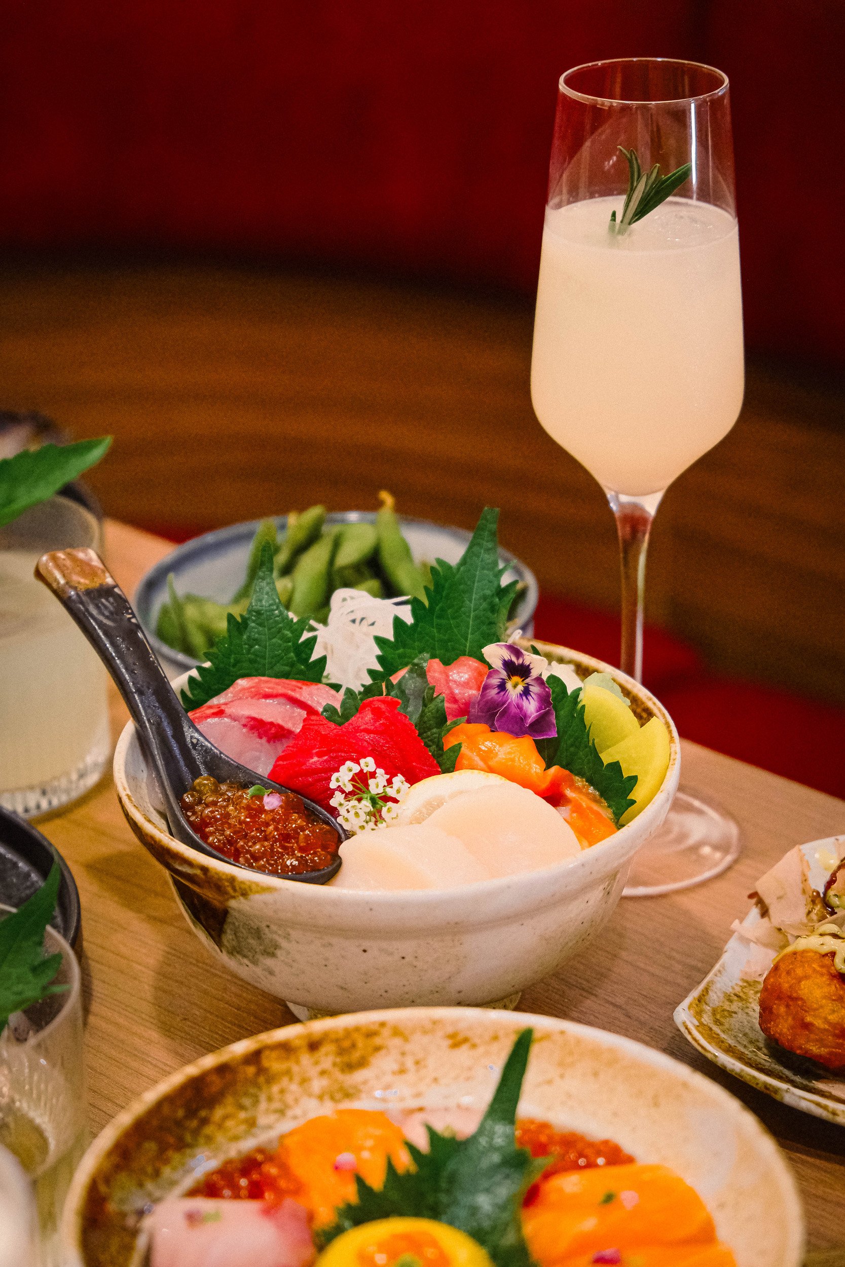 A bowl of colorful Japanese sashimi with various slices of fish, garnishes, and toppings, accompanied by a small spoon of ikura (salmon roe). In the background, a glass of champagne with a sprig of herbs inside, on a wooden table with other dishes an