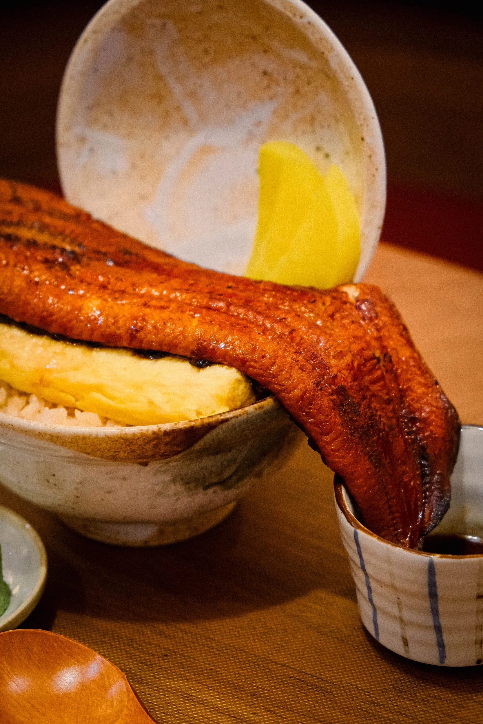 A Japanese meal featuring grilled eel with crispy skin, a bowl of rice with a cooked egg, and a wedge of lemon in a cup.