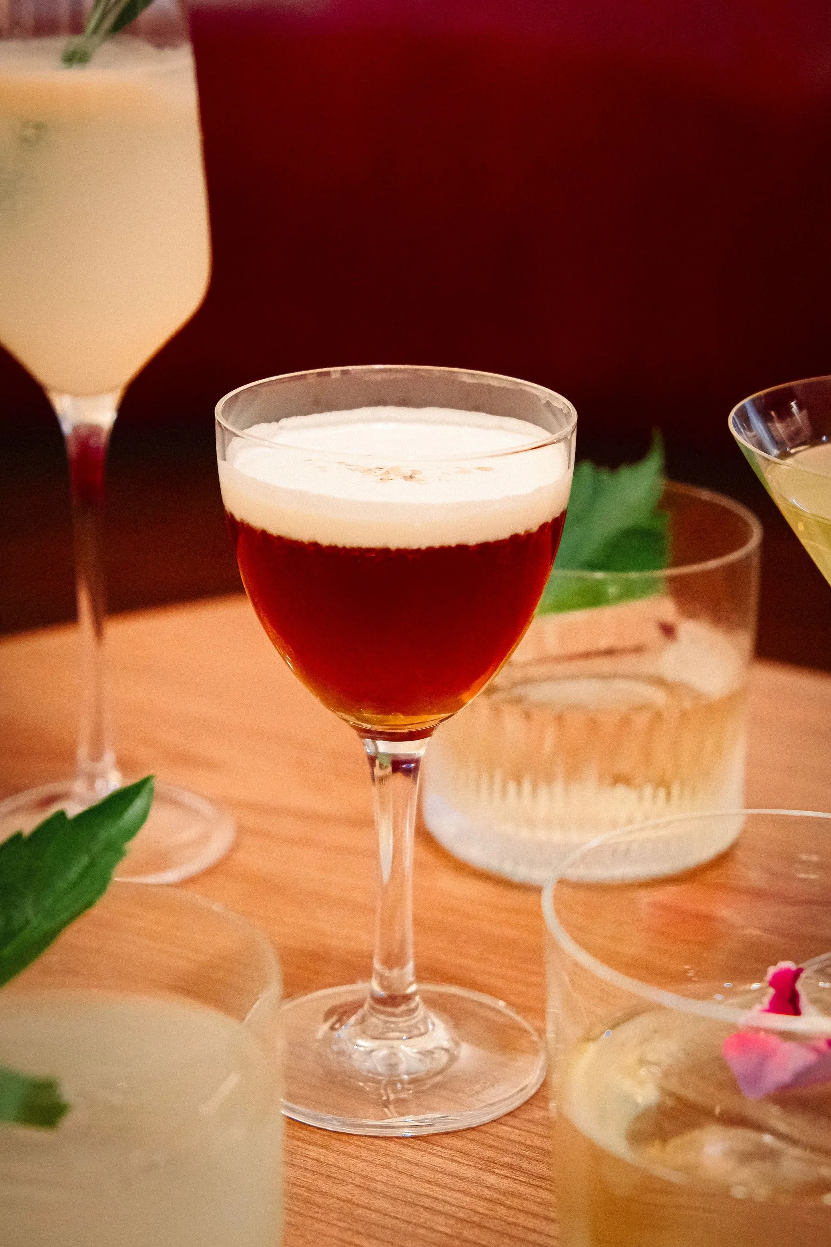 A close-up view of a red cocktail with a white foam layer on top in a martini glass, with other cocktail glasses and candles in the background.