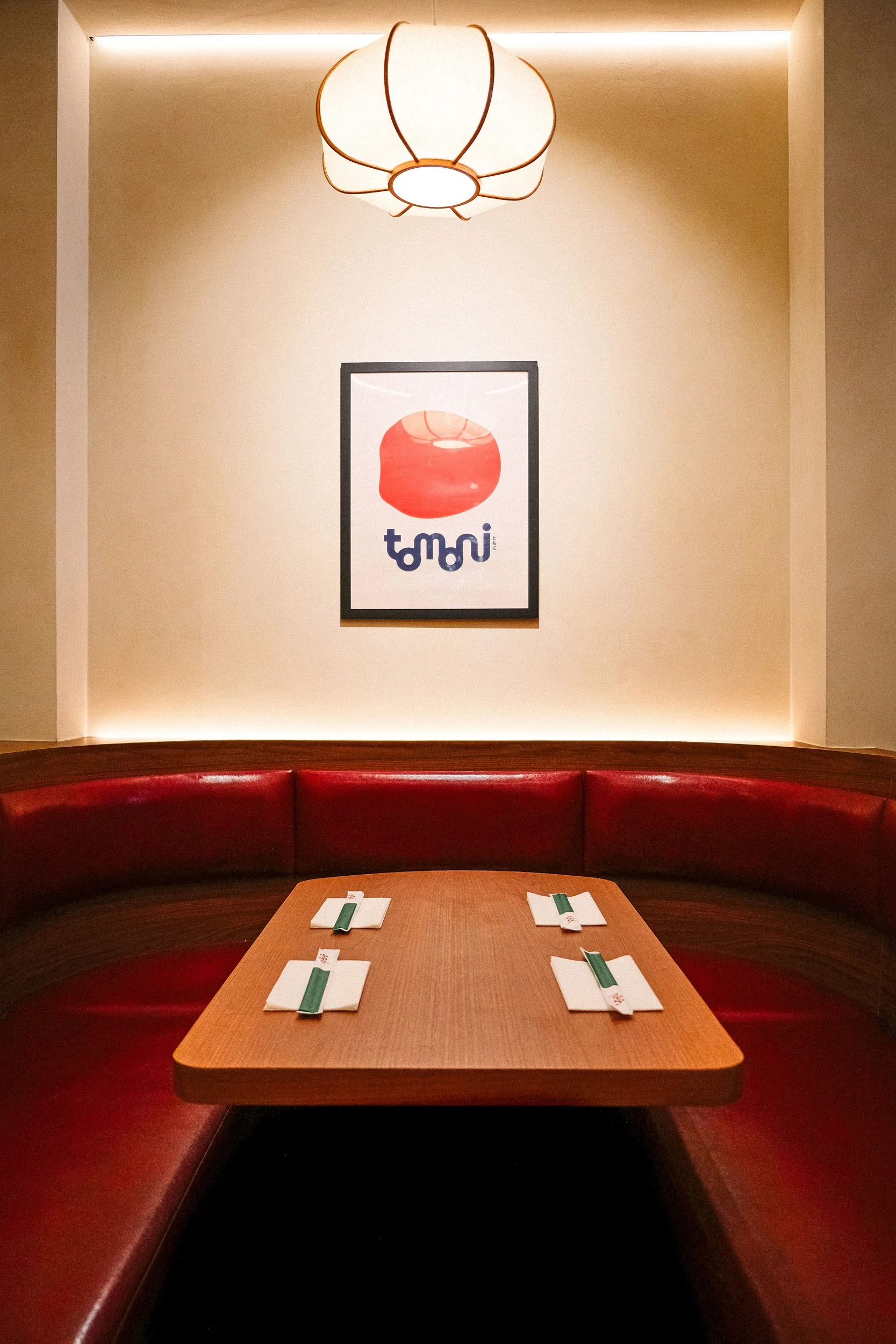 A restaurant dining area with a wooden table surrounded by a red curved booth seat. On the table are four sets of all-white napkins and green chopsticks. A framed artwork of a red lantern with Japanese characters is hanging on the beige wall behind t