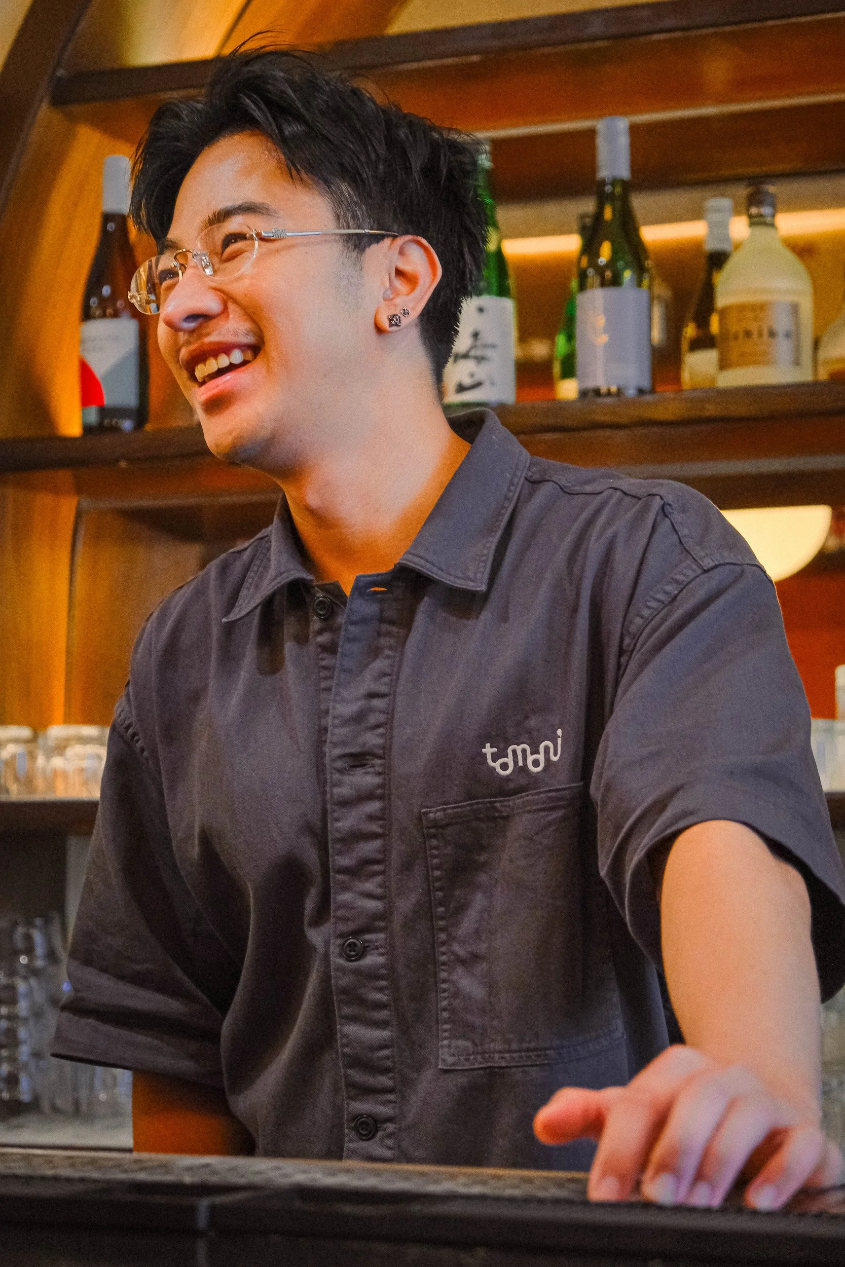 A young man with glasses and earrings wearing a dark button-up shirt smiling, with bottles of liquor on shelves in the background.