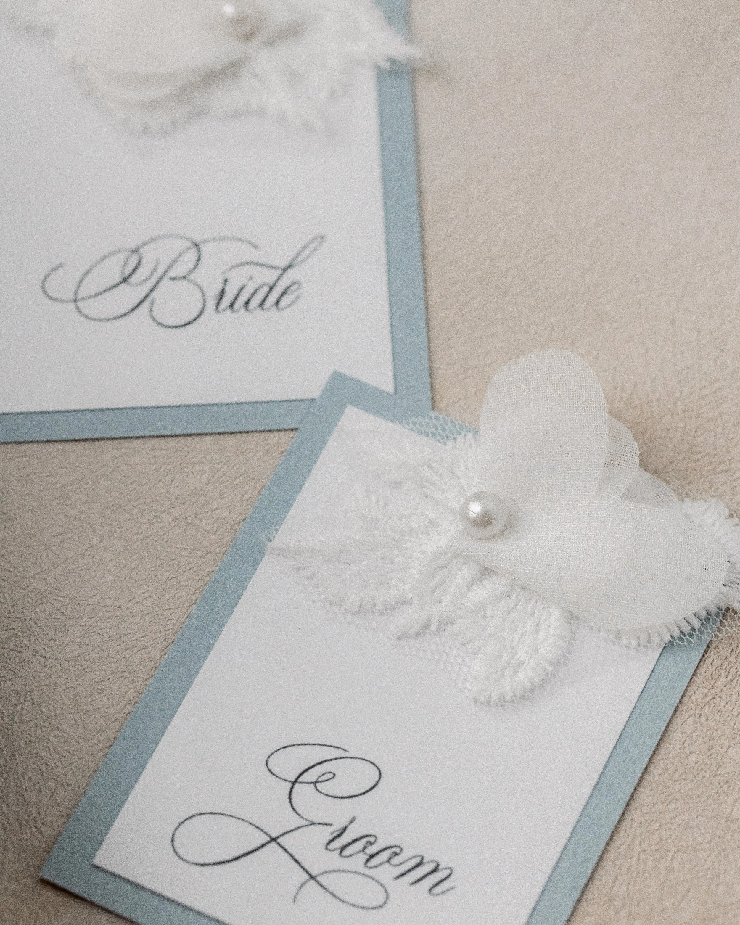 Bride and groom place cards echo the textures and tonal palette of the suite, integrating pearl-stitched flowers and subtle dimensional flourishes for a cohesive tablescape.