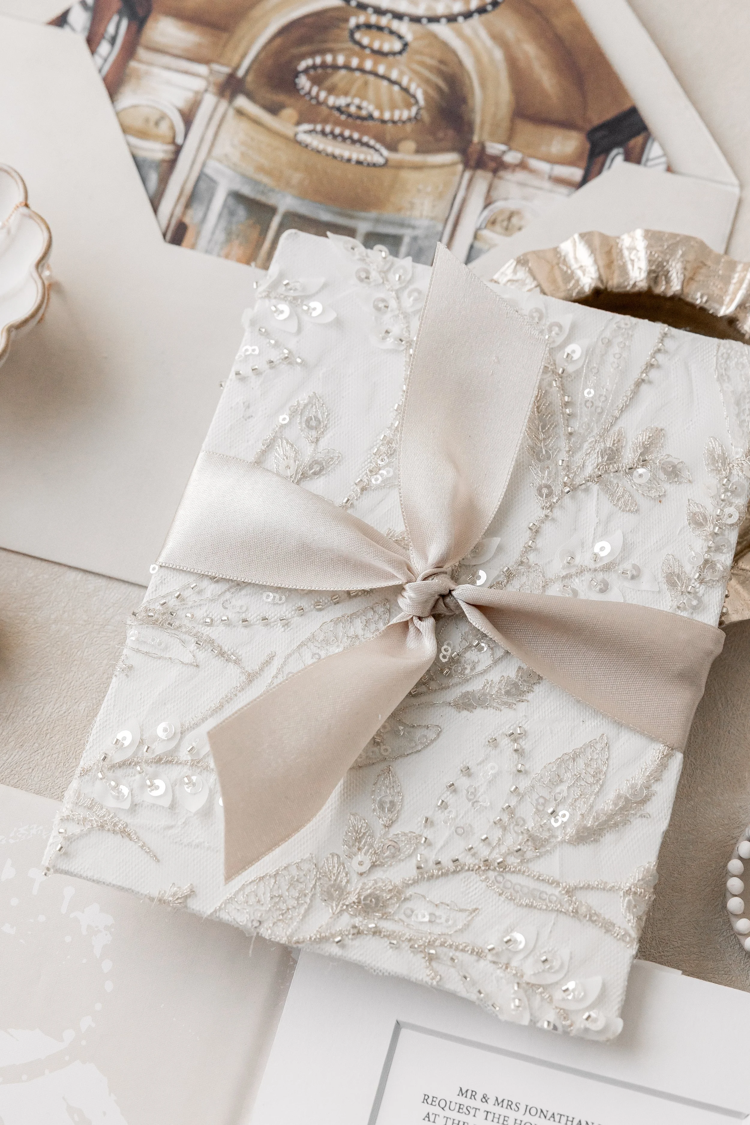 The signature book-style cover is wrapped in embroidered and sequined leaf detailing and finished with a champagne knot-tied ribbon, creating a sculptural first impression that feels both refined and romantic.