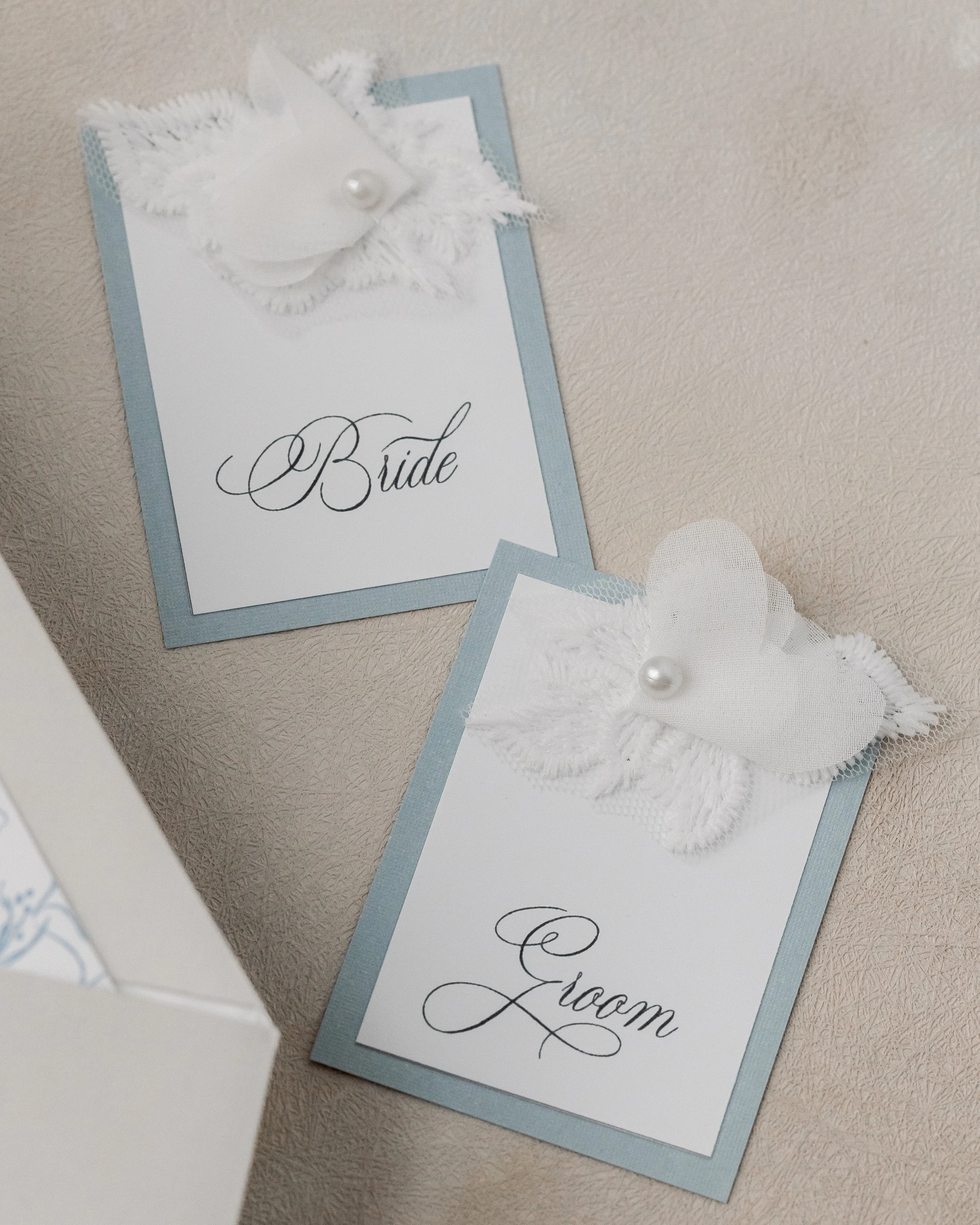 Dusty blue matte cards feature matching floral fabric elements and pearl accents, with “Bride” and “Groom” printed in coordinated script for a cohesive tablescape presentation.