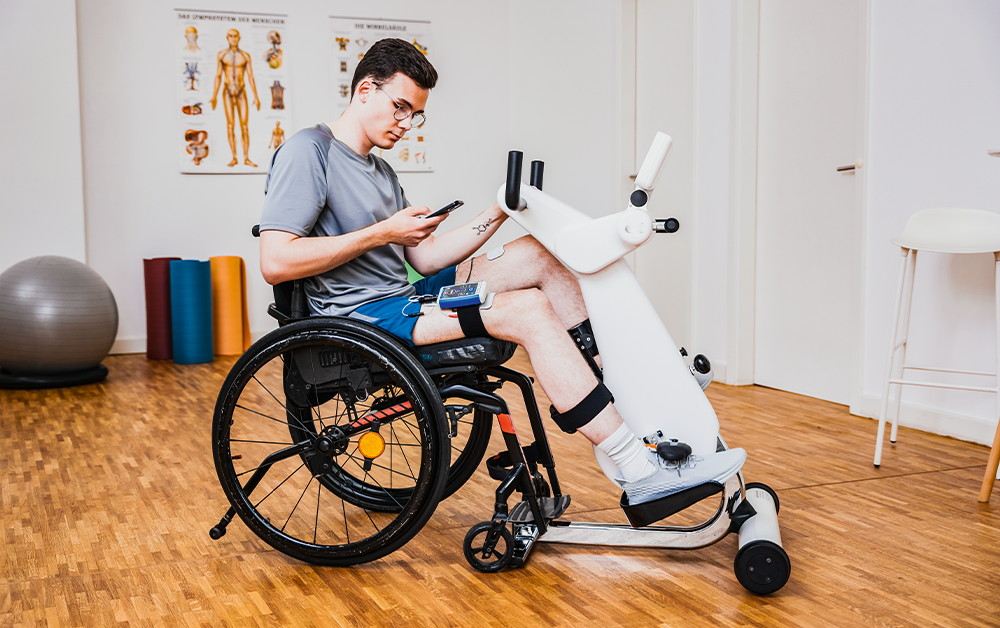 Person in a wheelchair using a robotic rehabilitation device on their legs in a therapy room.