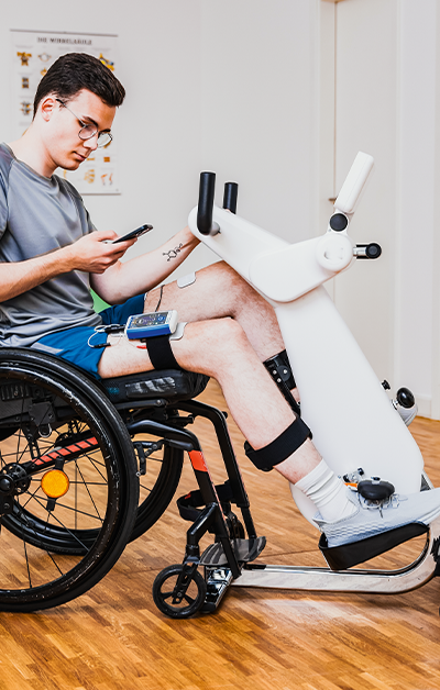 A person in a wheelchair using a robotic leg exercise machine while looking at a smartphone.