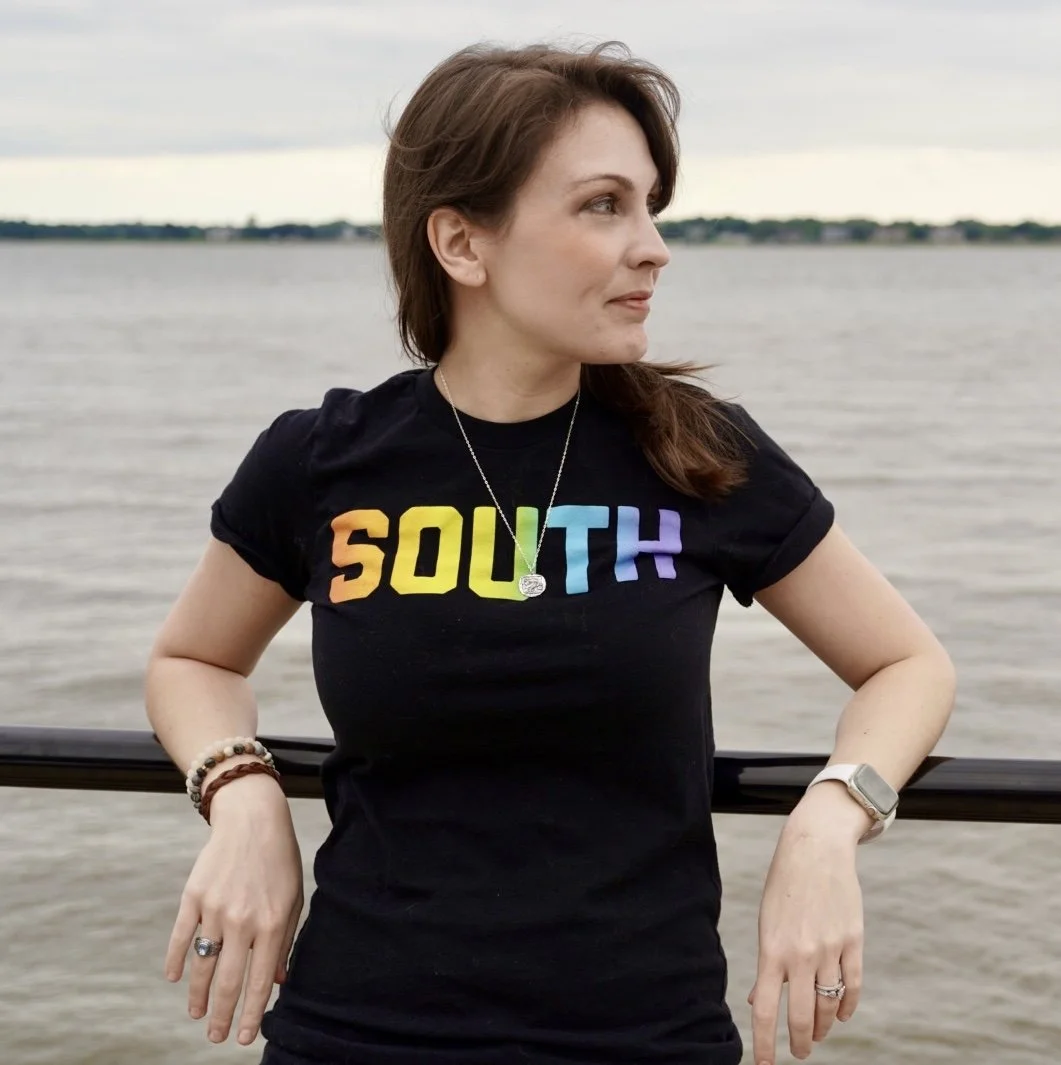 a photo of Kendra Winchester, a white person with long brown hair. She standing in front of a mental railing while looking off to the right. She's wearing a black t-shirt with the word South in a rainbow font
