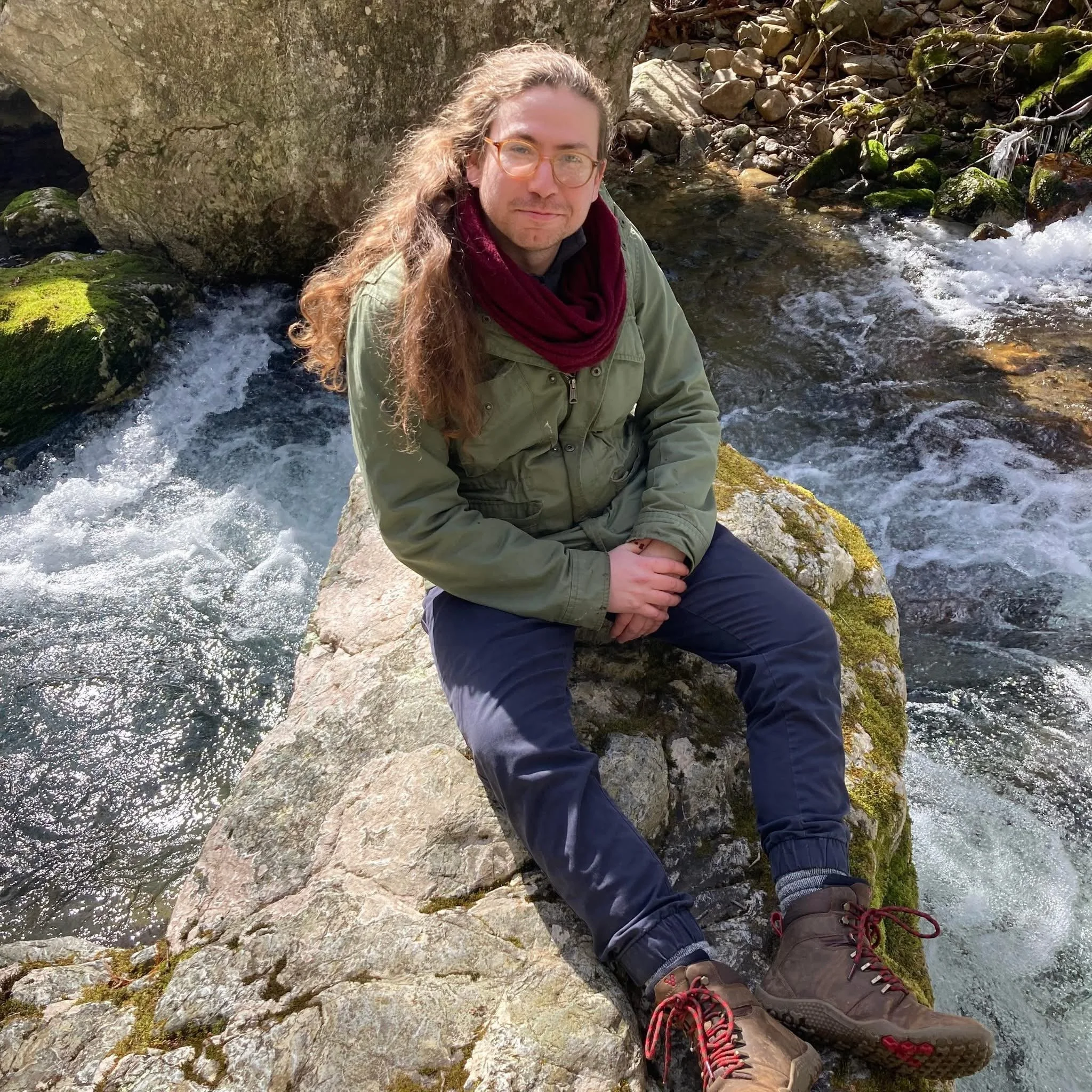 A photo of Davis Shoulders, a white person with long brown hair and glasses, wearing a green jacket, a maroon infinity scarf, blue pants, and brown boots sits on a boulder in the middle of a mountain stream smiling gently towards the photographer.