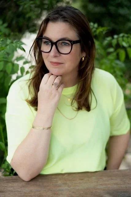 A woman with brown hair, glasses, and a yellow shirt sitting outdoors with greenery in the background.