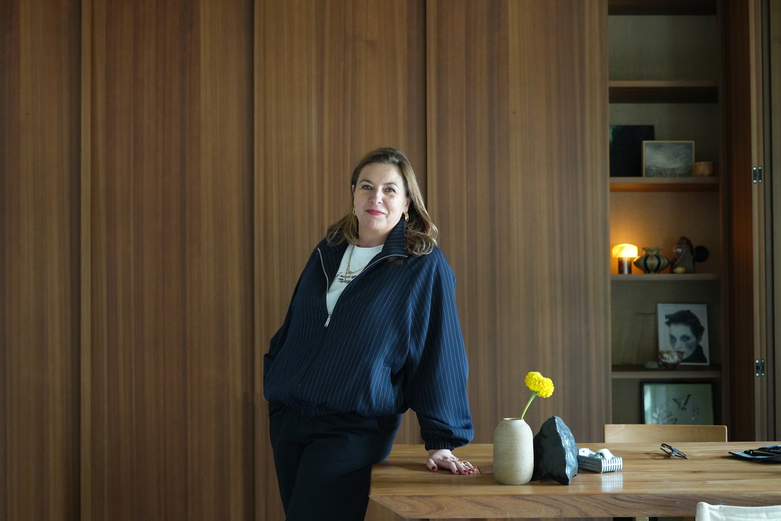 Liesbeth Demolder leaning on a wooden table with a yellow flower in a vase, in a warm wood-panelled room