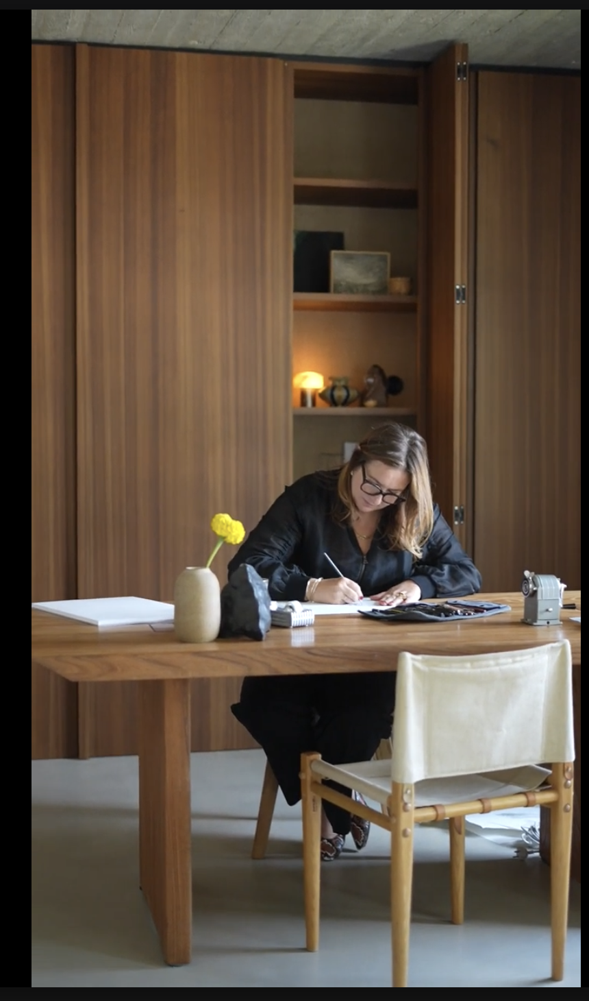 Liesbeth writing at a long wooden table, a single yellow flower in a vase beside her