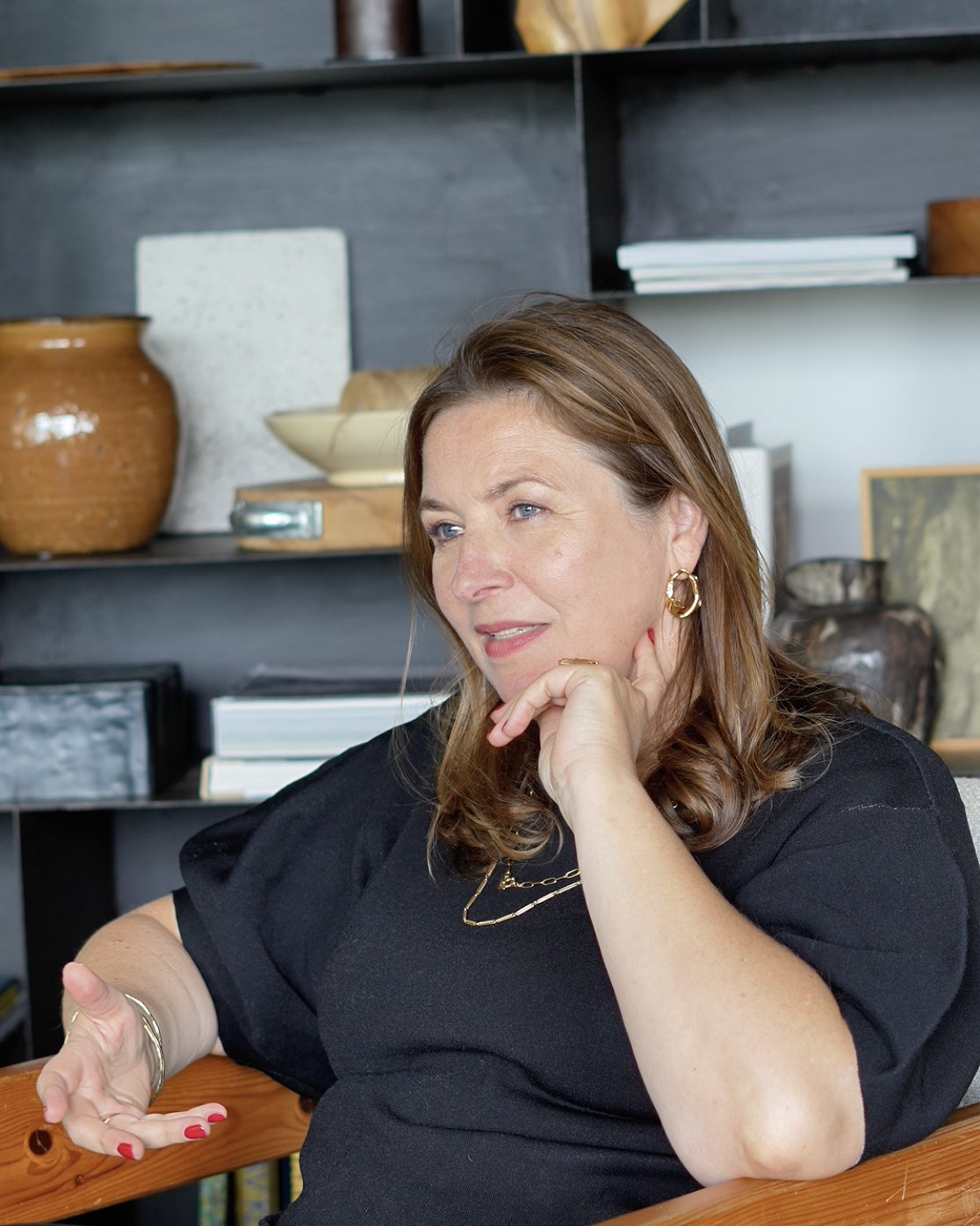 Liesbeth in conversation, seated in a wooden armchair in front of shelves with ceramics