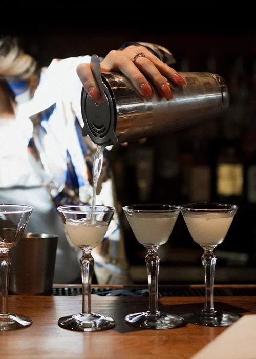 Person pouring a cocktail from a shaker into martini glasses at a bar.