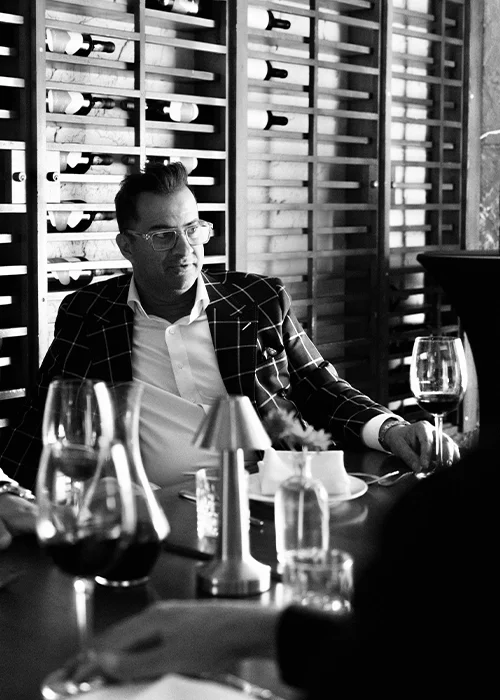 A man seated at a restaurant table with wine glasses, a water glass, and a salt shaker, in front of a wine rack wall.