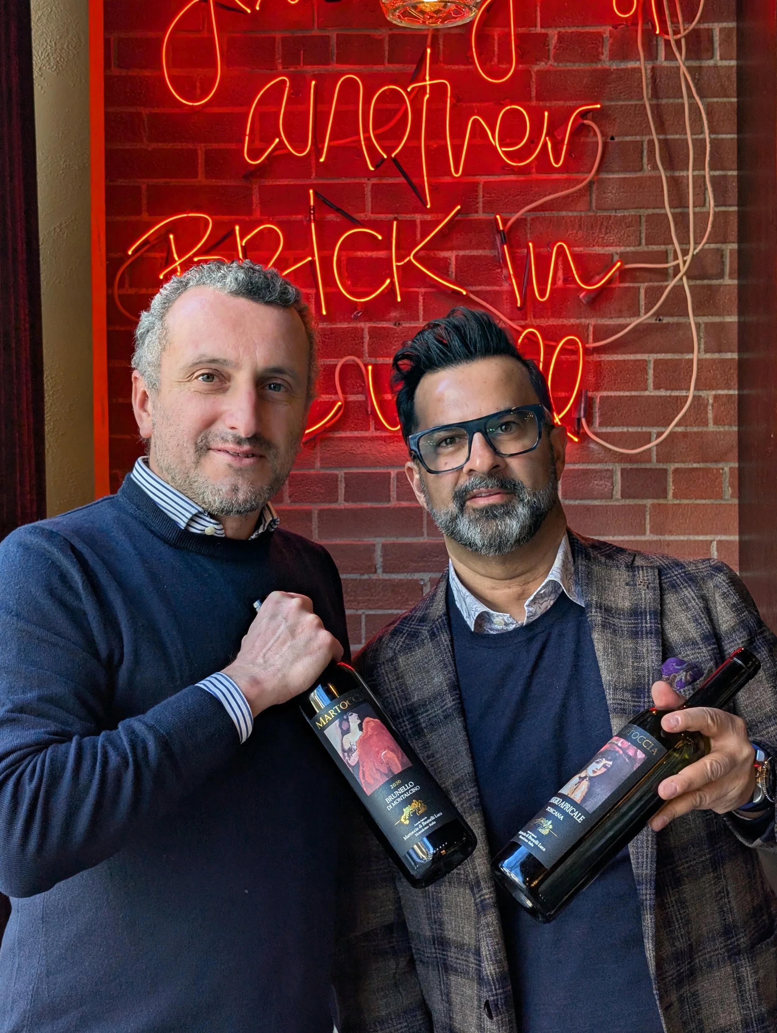 Two men standing in front of a brick wall with a neon sign that says 'another quick win'. The man on the left has light hair and beard, wearing a dark sweater over a striped shirt, holding a bottle of wine. The man on the right has dark hair, glasses