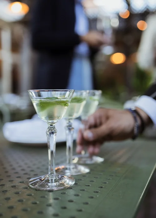 Two martini glasses filled with green cocktail garnished with lime, on a table at an outdoor setting, with people in the background.