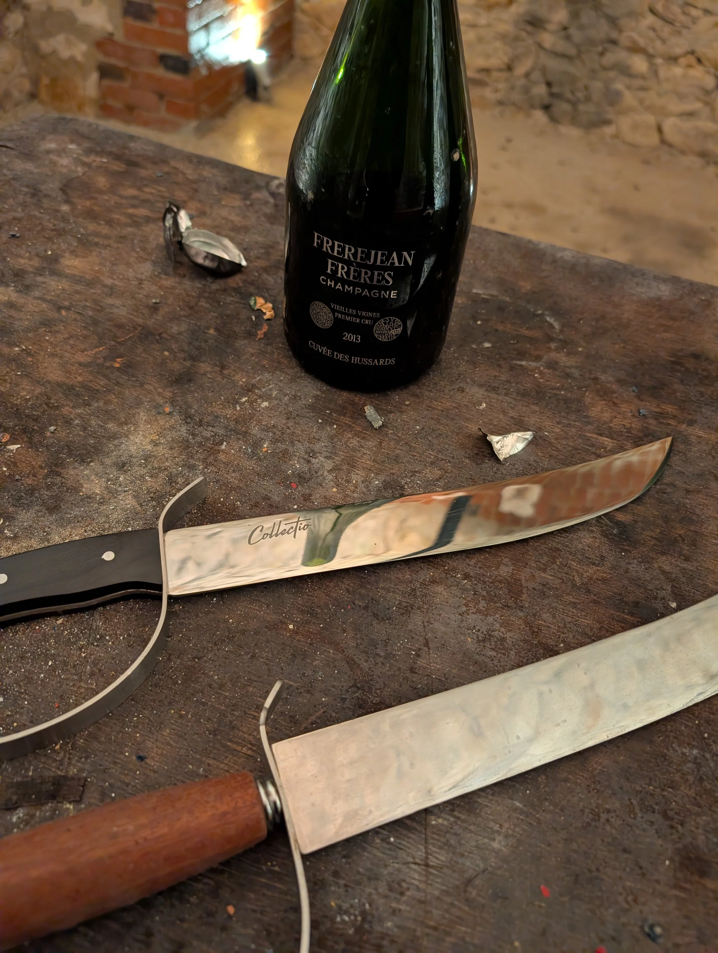 A wine bottle, a cheese knife, and a bread knife on a rustic wooden table. The wine is a 2013 Frerejean Freres Champagne. The cheese knife is labeled 'Collettio'.