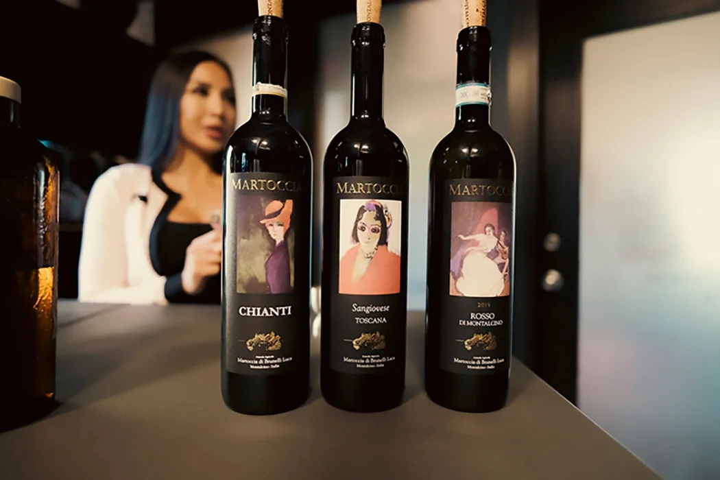 Three bottles of Martoccia wine with illustrated labels featuring women in various poses, placed on a counter with a blurred woman in the background.