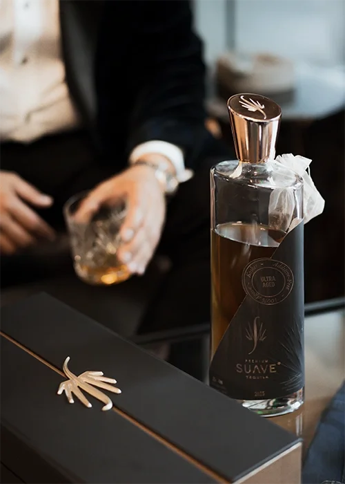 A bottle of tequila with a decorative gold leaf stopper on top, placed on a black surface. In the background, a person in a dark suit and white shirt is holding a glass with an amber-colored drink and ice. A black box with a gold x-shaped decoration 