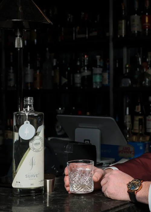 A bartender pours a clear liquid from a bottle labeled 'Suave' into a glass with a textured pattern, on a dark bar counter, with liquor bottles in the background.