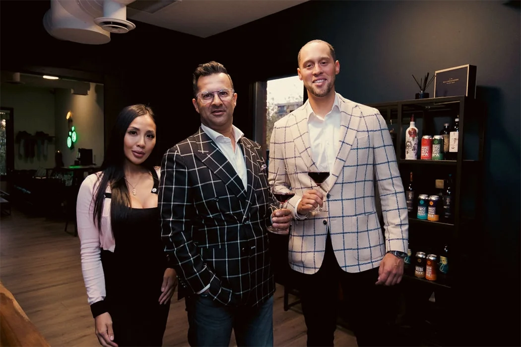 Three people, two men and one woman, stand indoors holding drinks, with shelves of beverages and a window in the background.