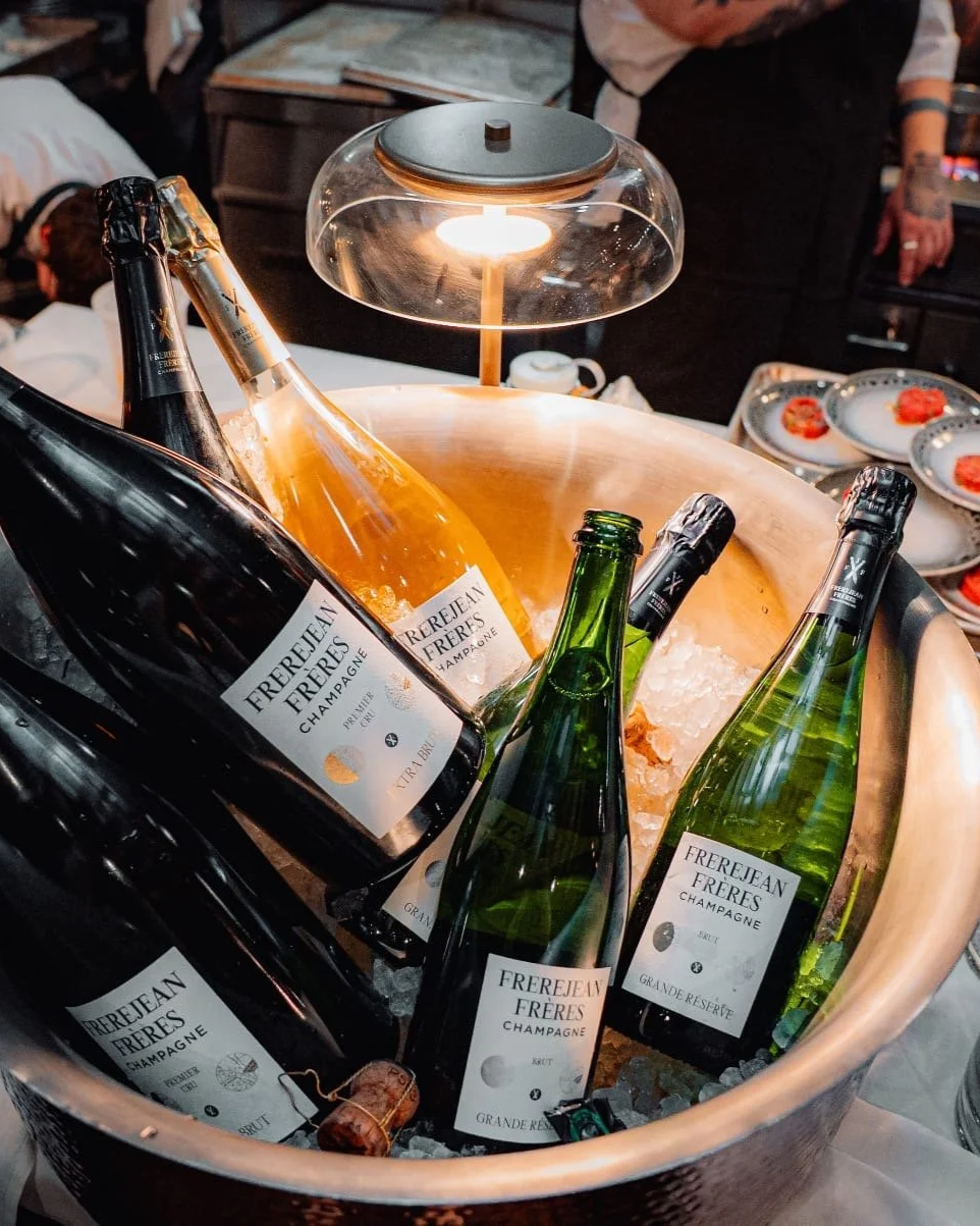 Bottles of Frejean Frères champagne chilling in an ice bucket with a lamp overhead and plates of food in the background.