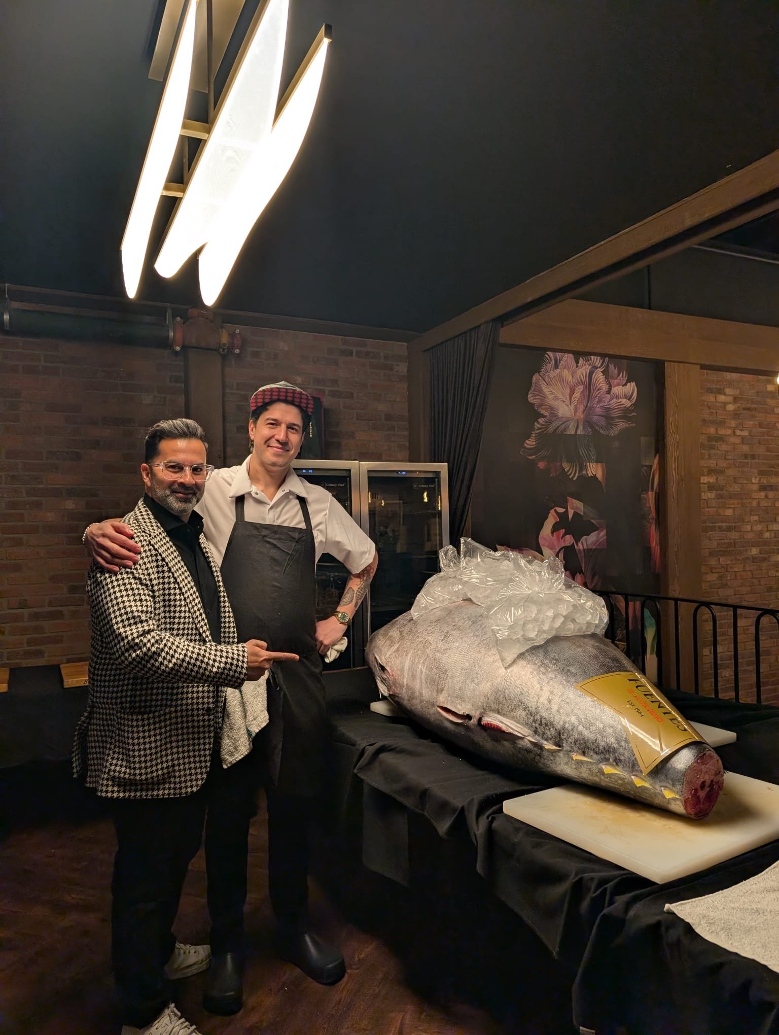 Two men standing next to a large fish on a table in a restaurant.