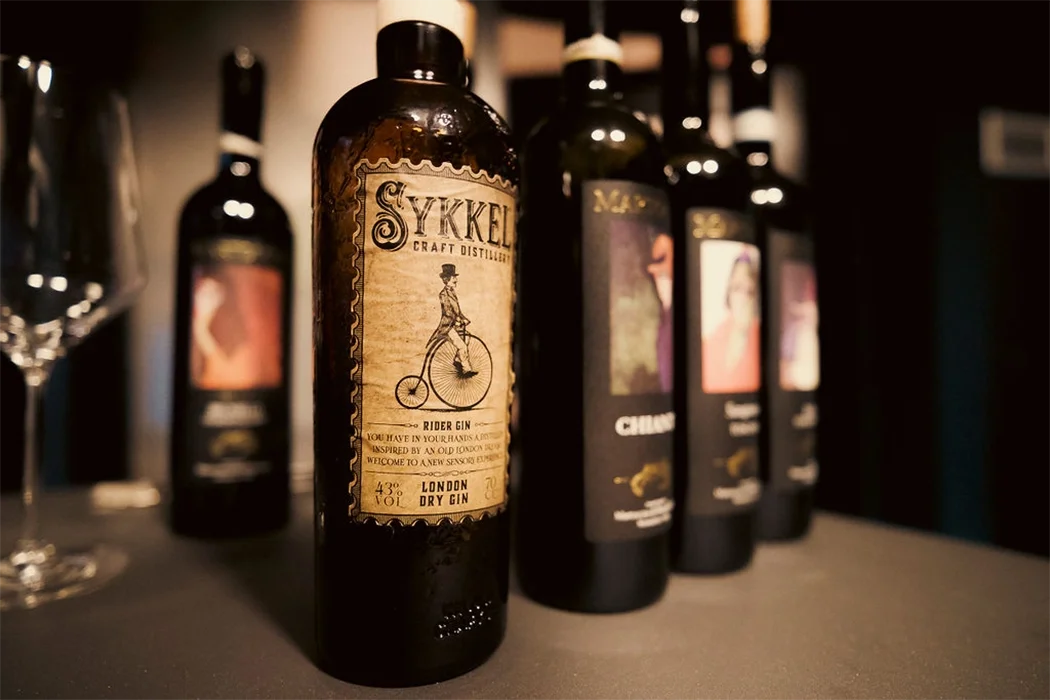 Bottles of craft gin lined up on a dark surface, with the front bottle labeled "sykkel" rider gin featuring vintage-style label art of a man riding a penny-farthing bicycle, and other bottles blurred in the background.