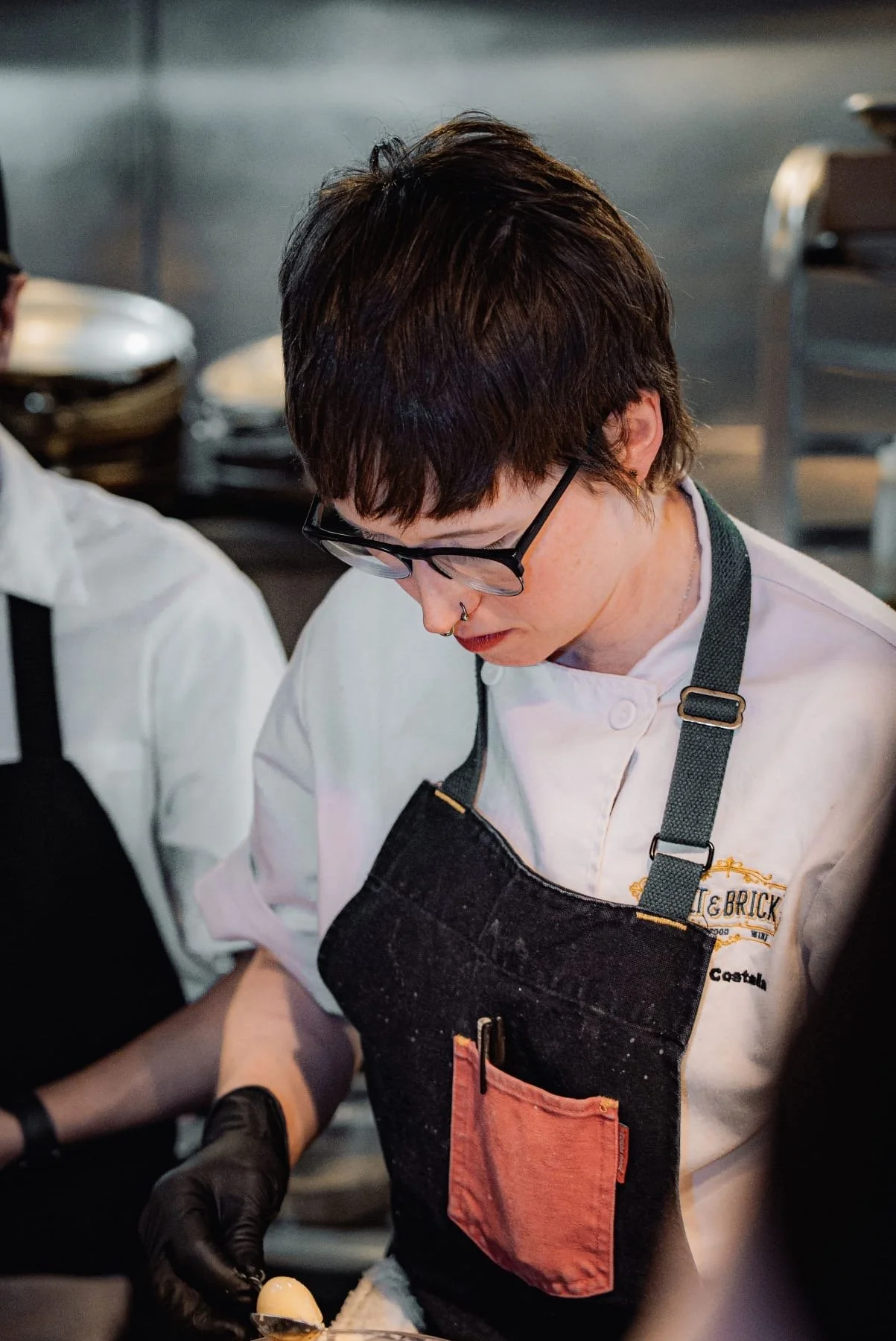 A person with short dark hair, glasses, and a septum piercing, wearing a white chef jacket and a black apron with a pink pocket, appears to be working in a professional kitchen.