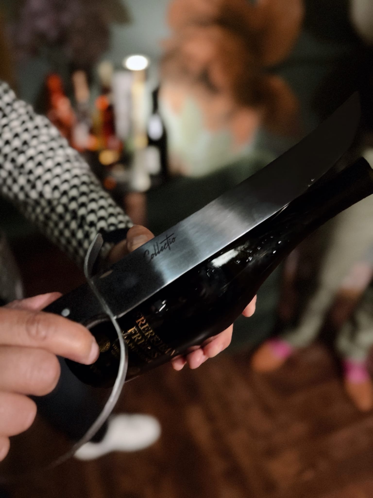 Person holding a large chef's knife over a dark bottle, with a blurred background of people and a bar or restaurant setting.