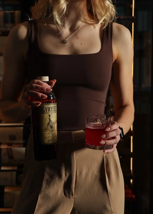 A woman holding a bottle of liquor and a glass of red drink in a dimly lit setting.