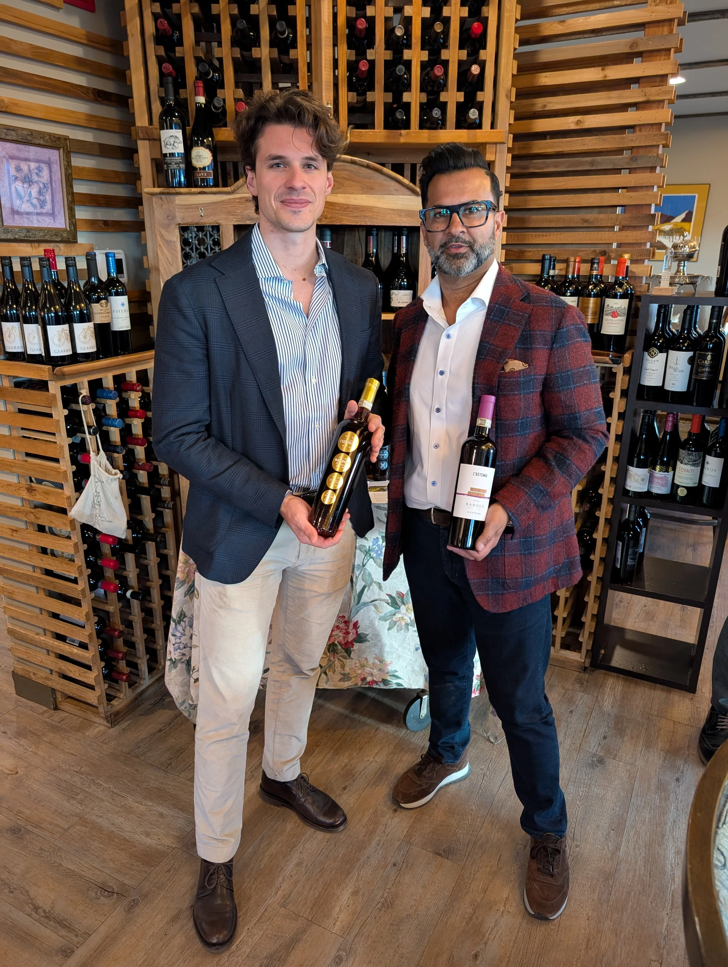 Two men standing in a wine shop, holding bottles of wine, with wine racks behind them.