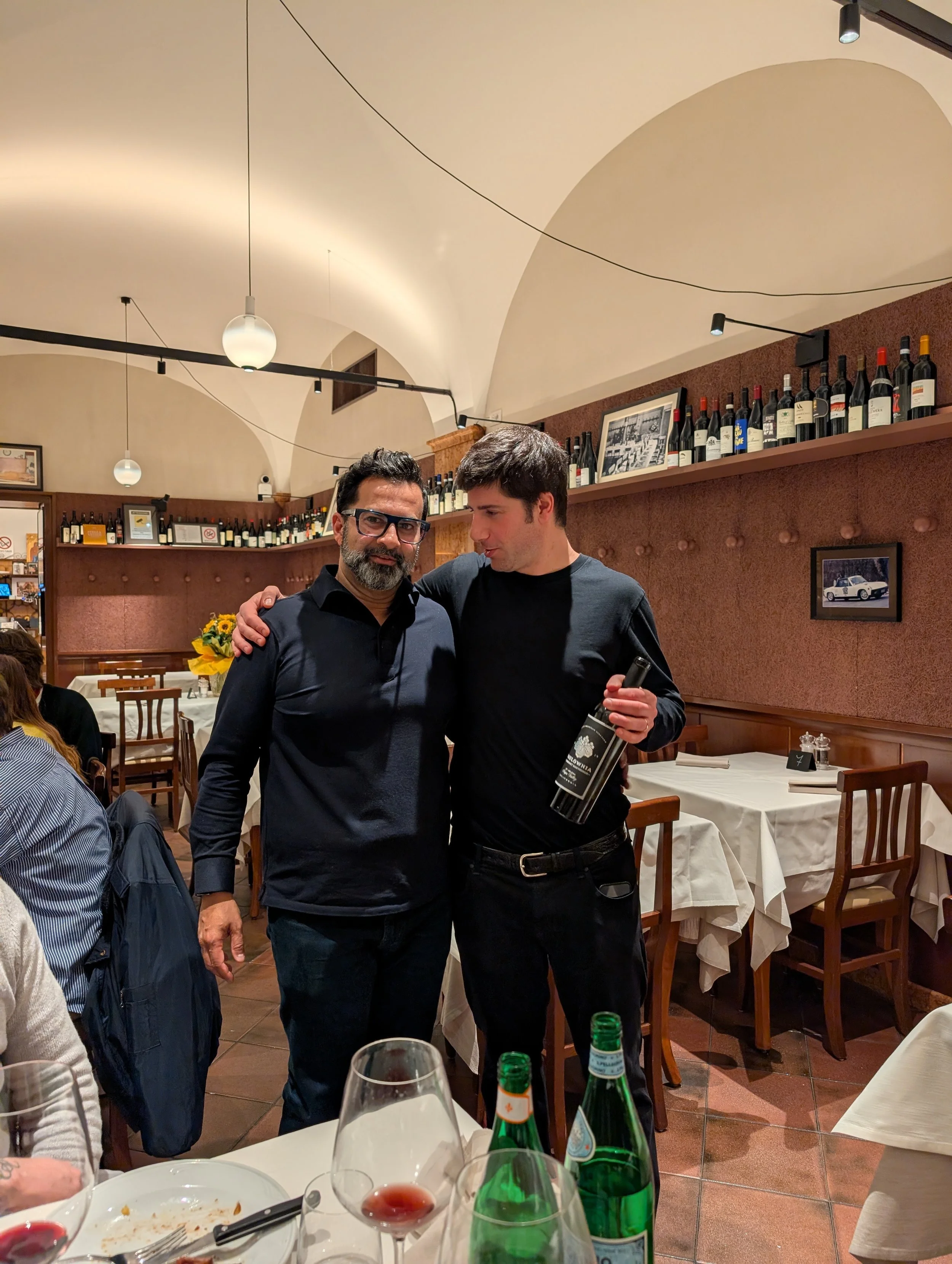 Two men standing together in a restaurant, one with glasses and a beard, the other holding a bottle of wine, both smiling and wrapping their arms around each other.